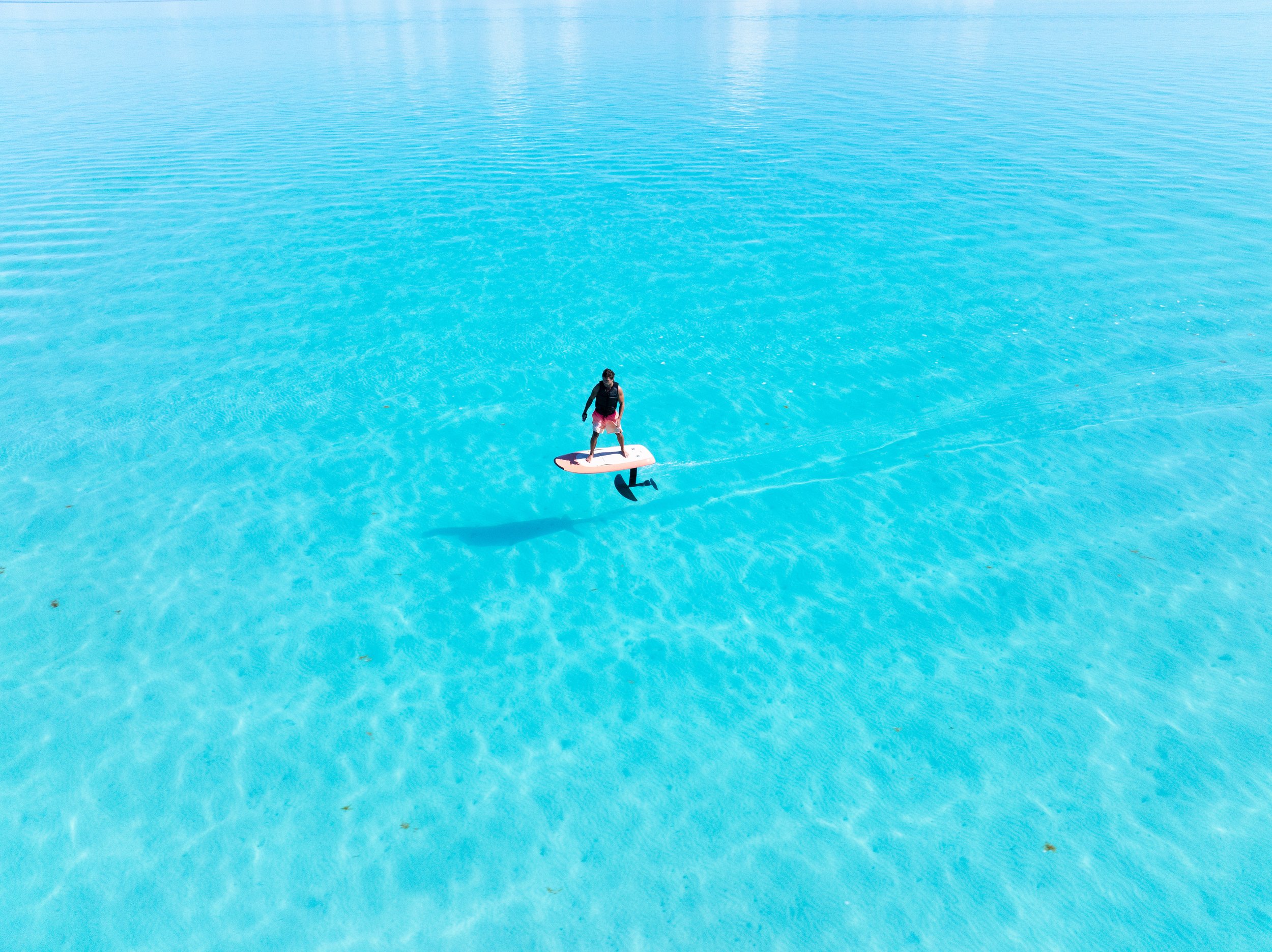 A person stands on an efoil on clear turquoise water in a tropical setting. Perfect for wakesurfing, wakeboarding, waterskiing, tubing. Located in Gracebay, Providenciales, Turks & Caicos.
