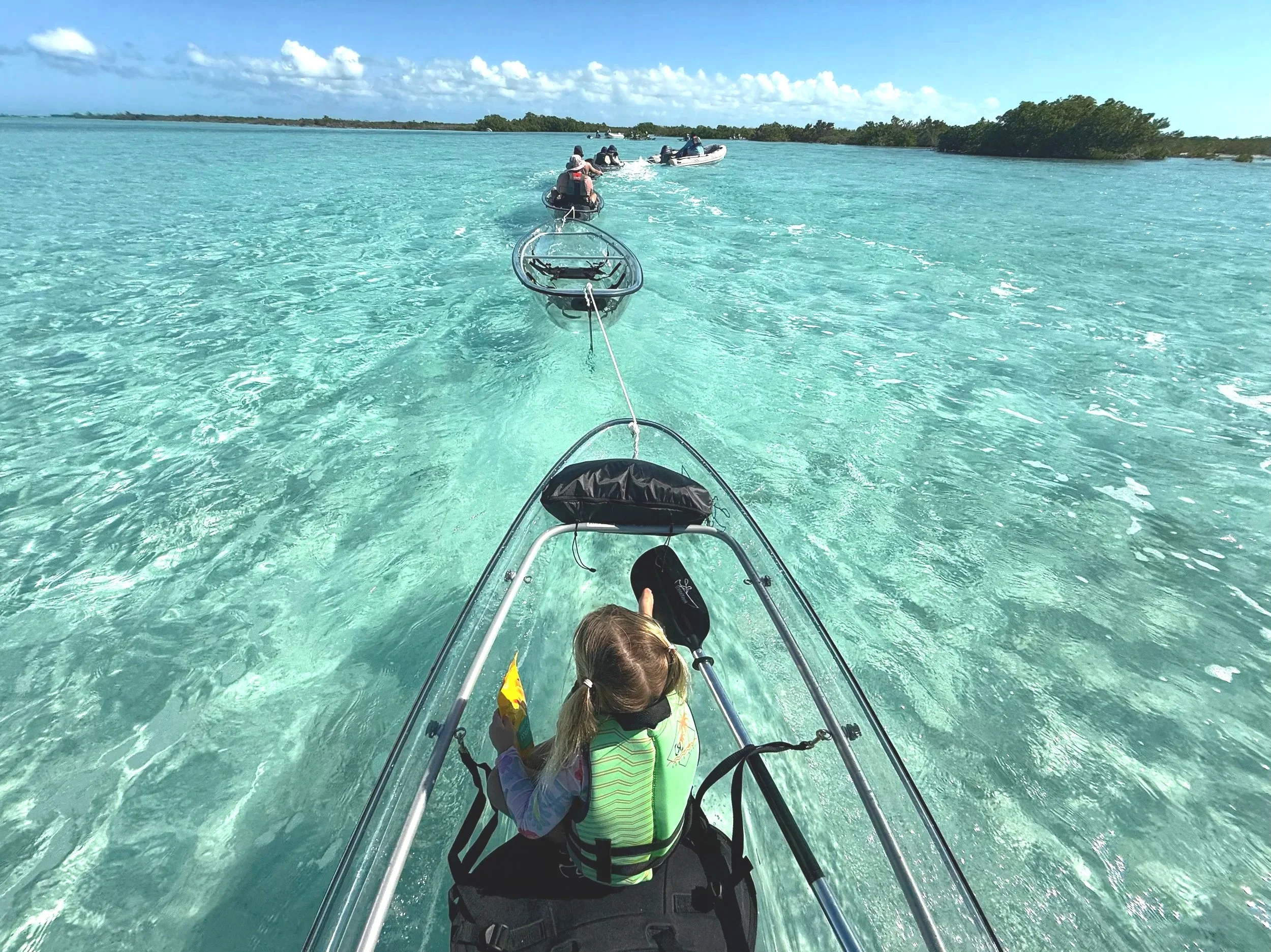 A group of people and children being towed in clear bottom kayak on clear, turquoise water near small islands under a partly cloudy sky. Searching for turtles, located in providenciales Turks & Caicos.