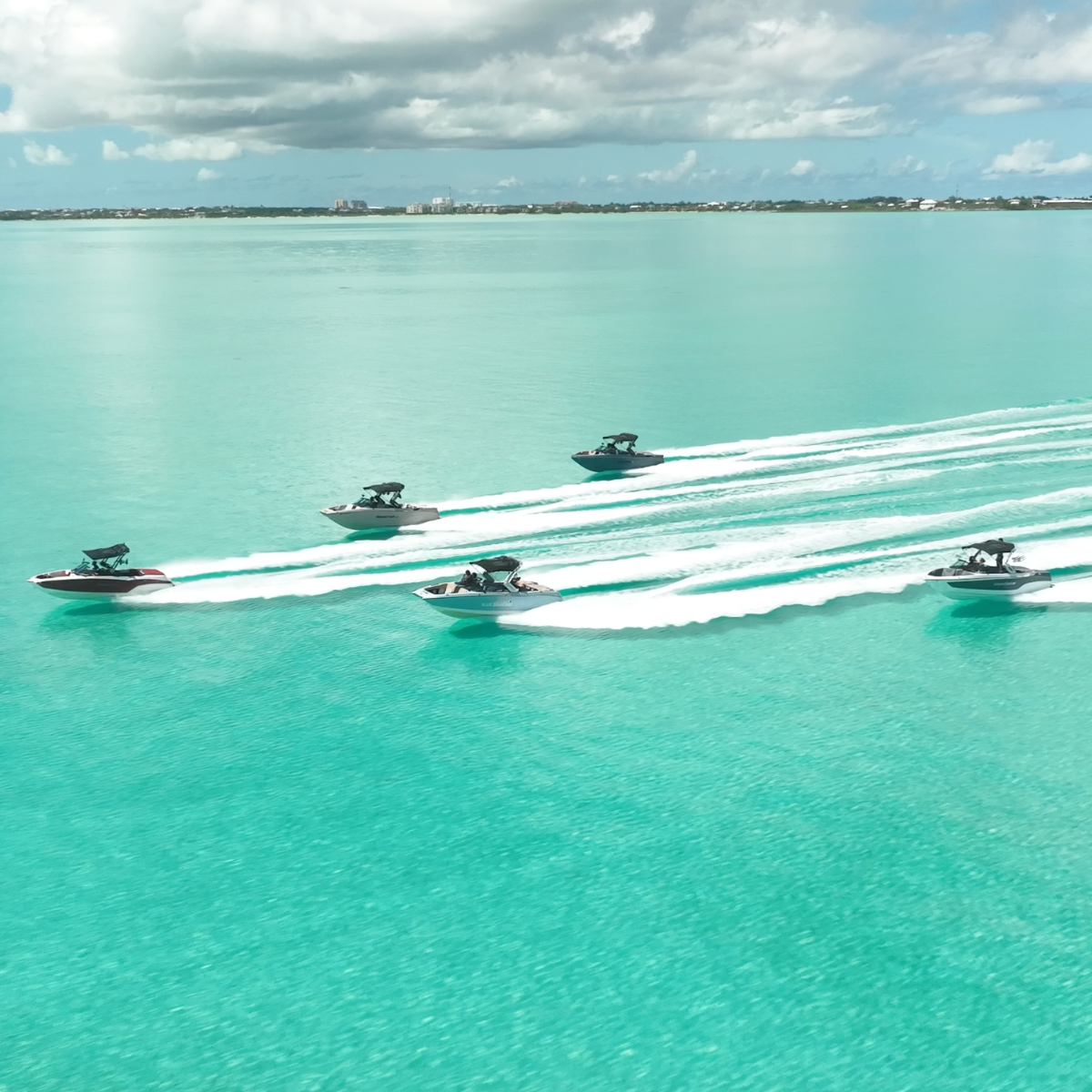 Five wake boats speeding across turquoise water, leaving white trails behind them, with an island skyline and sunny sky in the background. Perfect for wakesurfing, wakeboarding, waterskiing, tubing. Located in Gracebay, Providenciales, Turks & Caicos