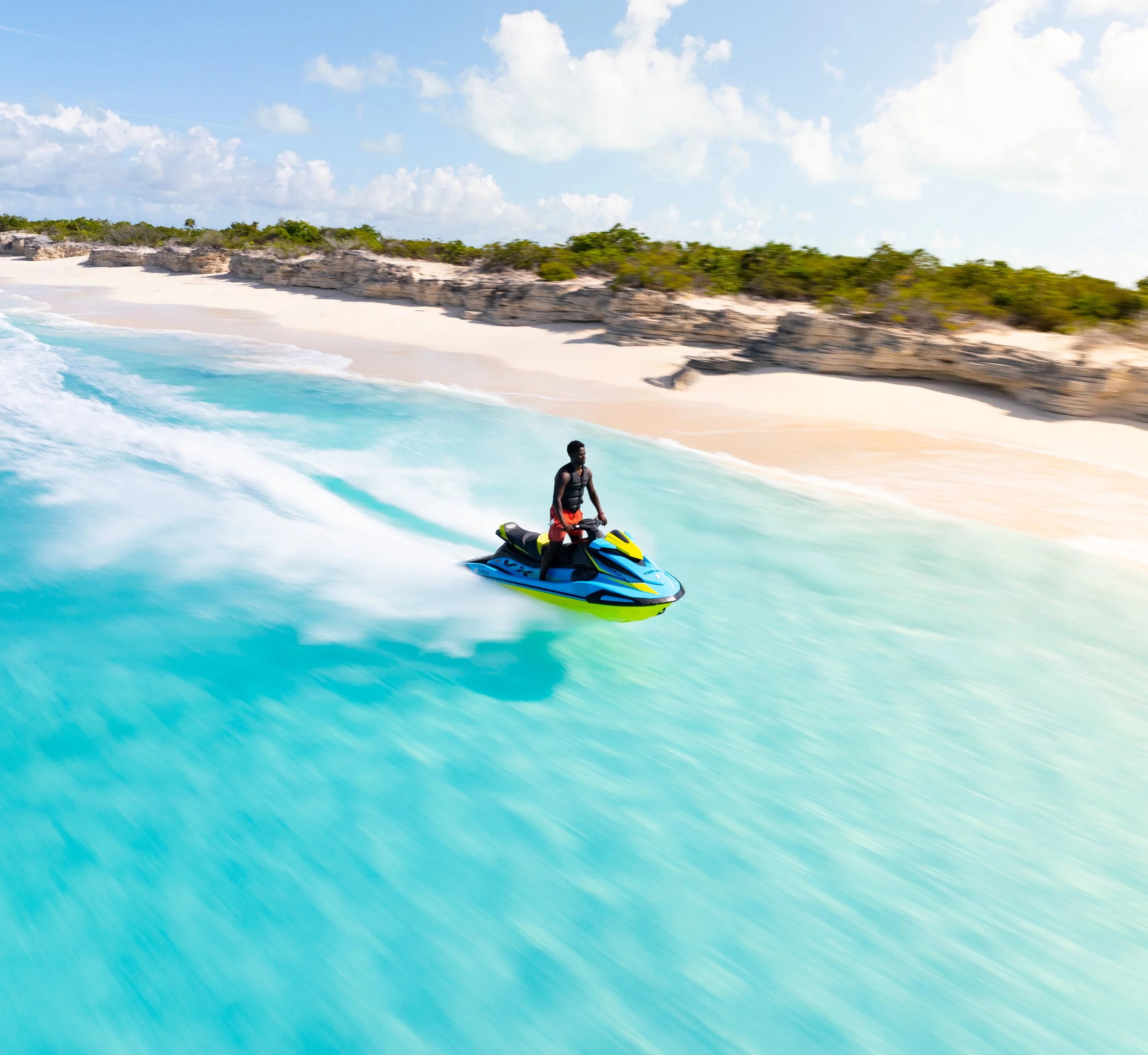 A man riding a jet ski on the turquoise ocean near a sandy beach with rock formations and green foliage, under a partly cloudy sky. Located in Gracebay, Providenciales, Turks & Caicos.