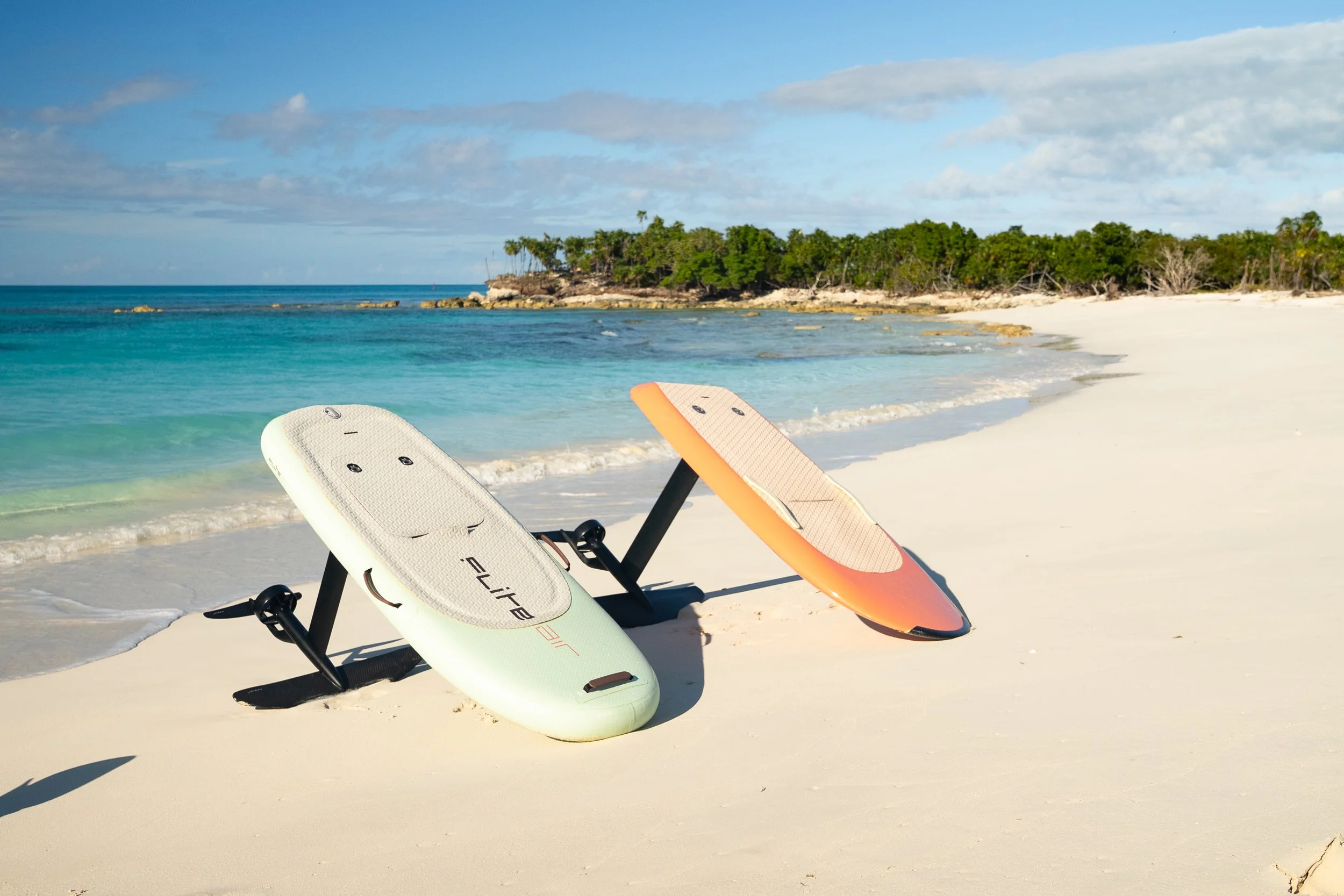 Two efoils, one white and one orange, resting on a sandy beach near the shoreline with a clear turquoise ocean and a green, tree-covered headland in the background. Located in Gracebay Turks & Caicos.