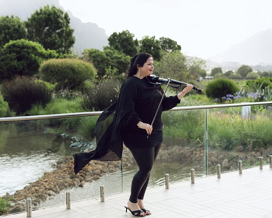 A woman with long dark hair and earrings, dressed in black with a flowing black cape, is playing a violin outdoors on a balcony overlooking a lush green garden and water.