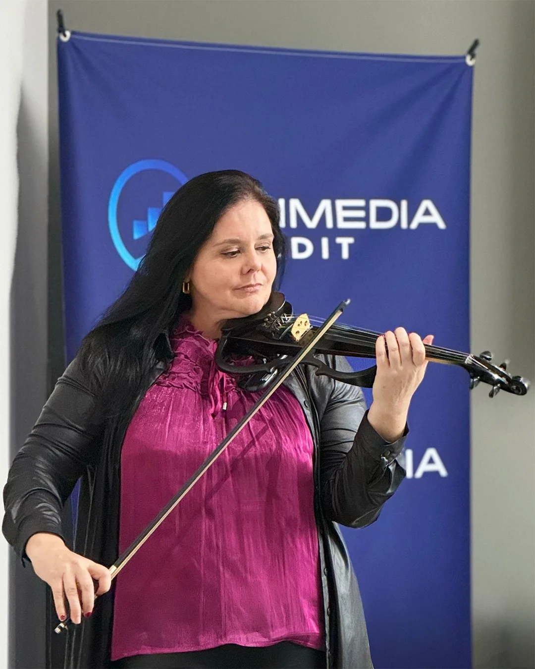 A woman playing the violin at a media event, standing in front of a blue backdrop with 'MEDIA' visible.