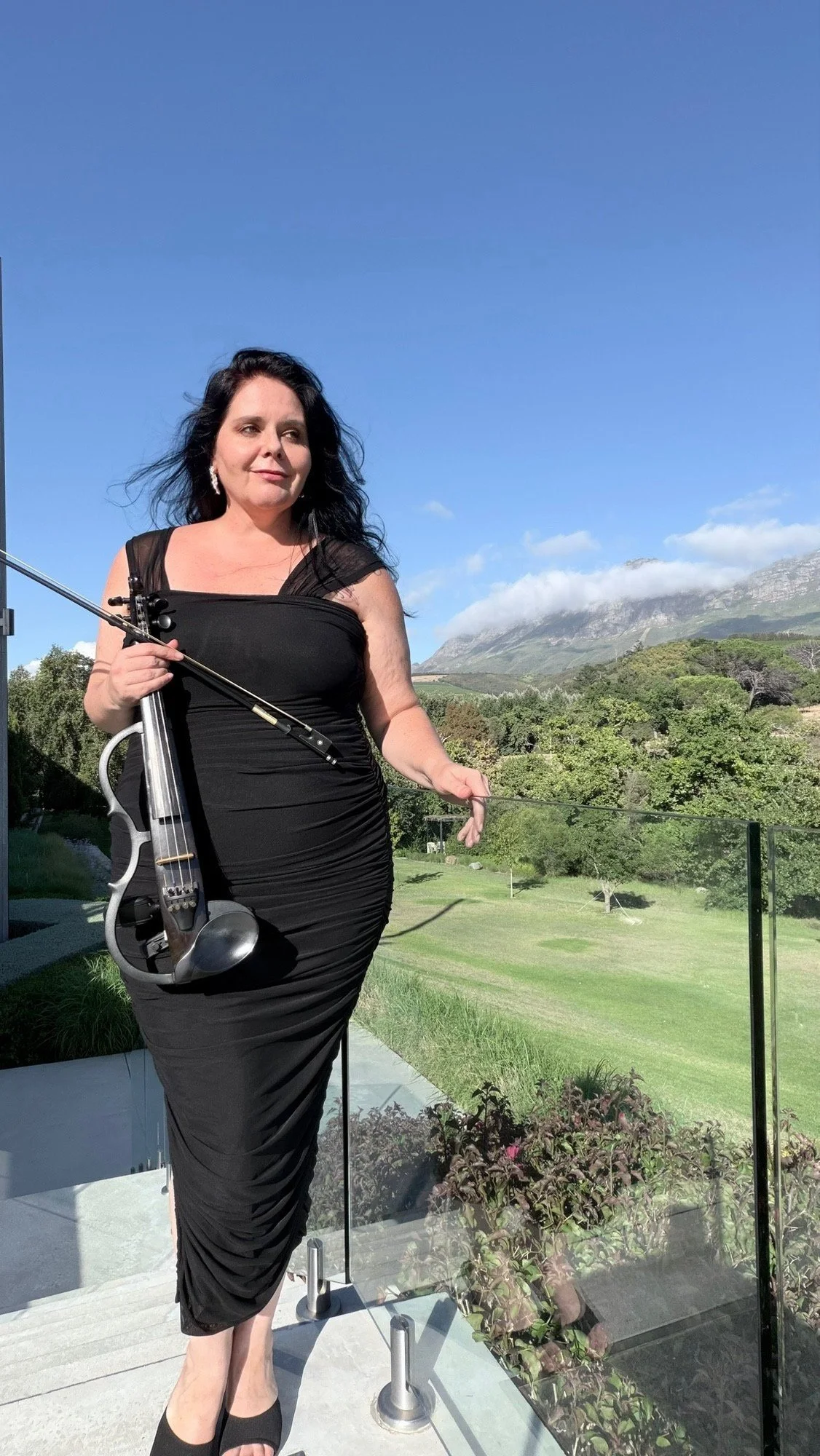 A woman in a black dress holding a violin outside on a balcony with scenic mountains and greenery in the background.