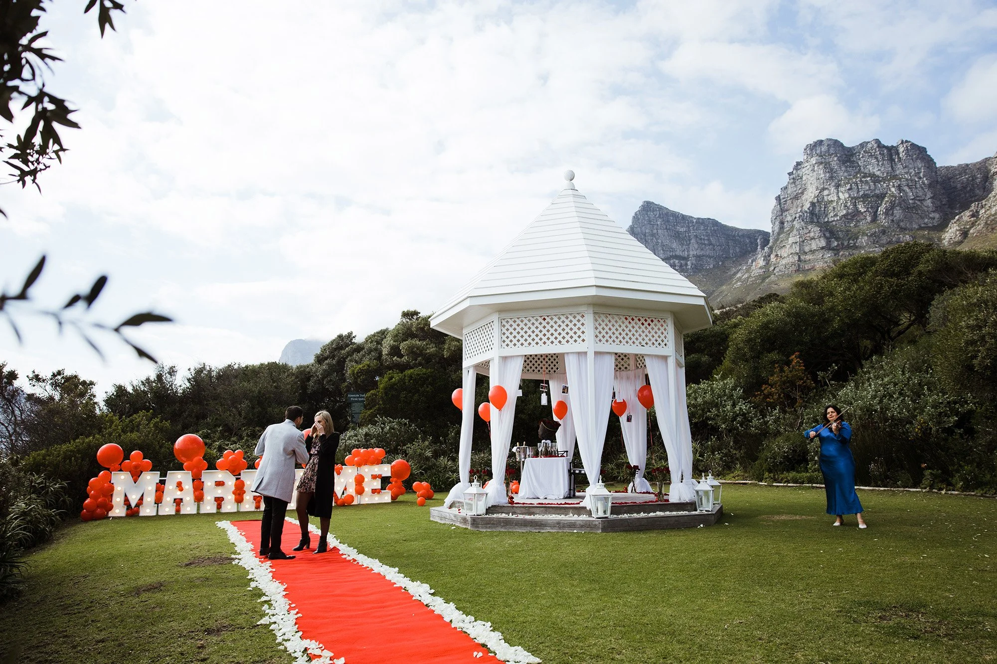 Larah Eksteen ready for a Proposal setup with a white gazebo decorated with white curtains and lanterns, red balloons, and a red aisle runner on green grass, with mountains and cloudy sky in the background.