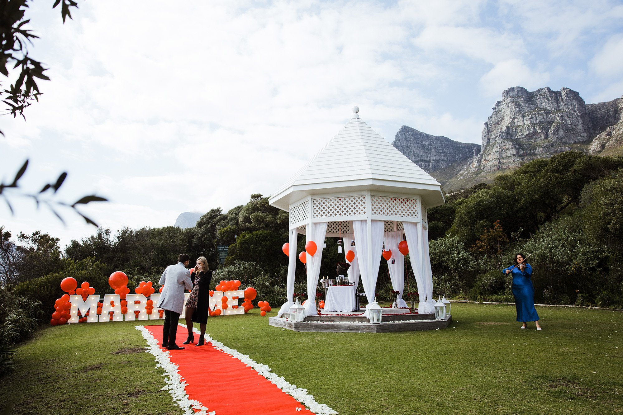 Larah Eksteen ready for a Proposal setup with a white gazebo decorated with white curtains and lanterns, red balloons, and a red aisle runner on green grass, with mountains and cloudy sky in the background.