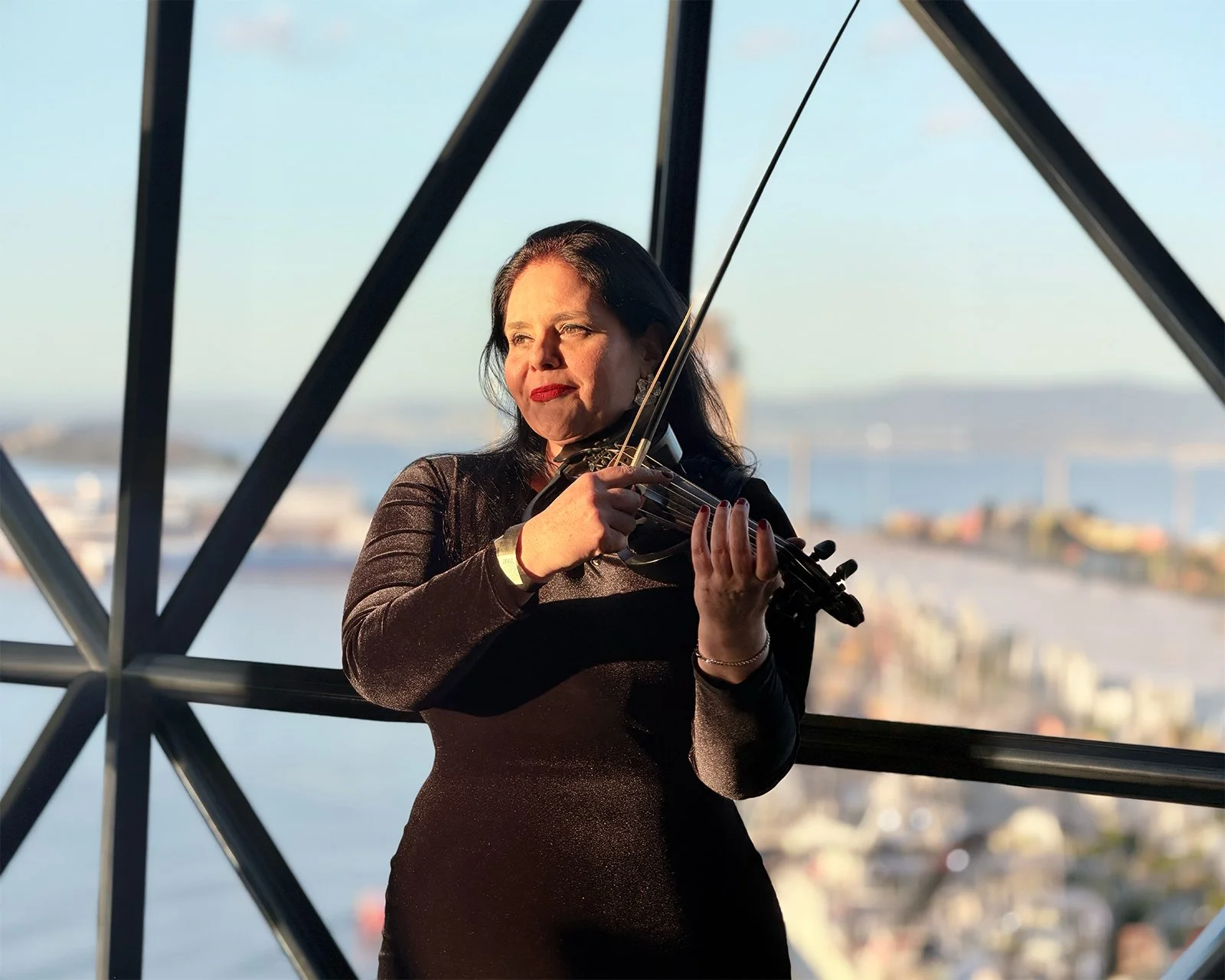 A woman playing the violin outdoors during sunset with a cityscape and water in the background.