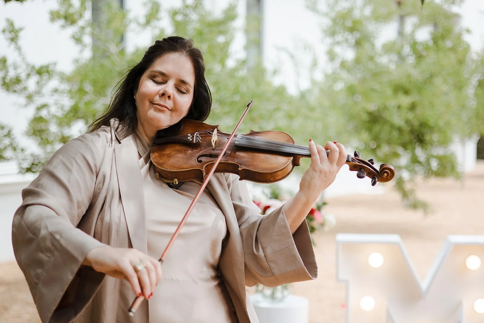 A woman playing the violin outdoors, with closed eyes and a serene expression, surrounded by greenery.