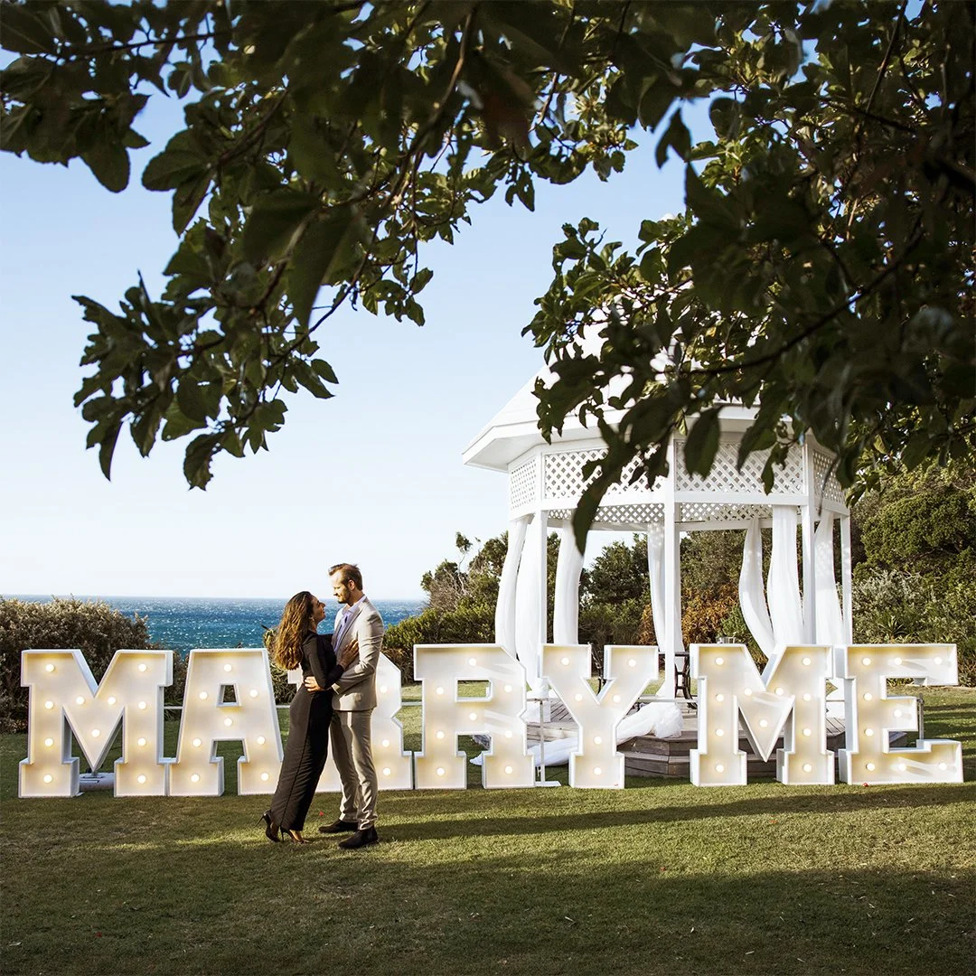 A couple stands close together in front of large, illuminated letters spelling "MARRY ME" on a green lawn, with a white gazebo and trees in the background, and the ocean visible in the distance under a clear blue sky.