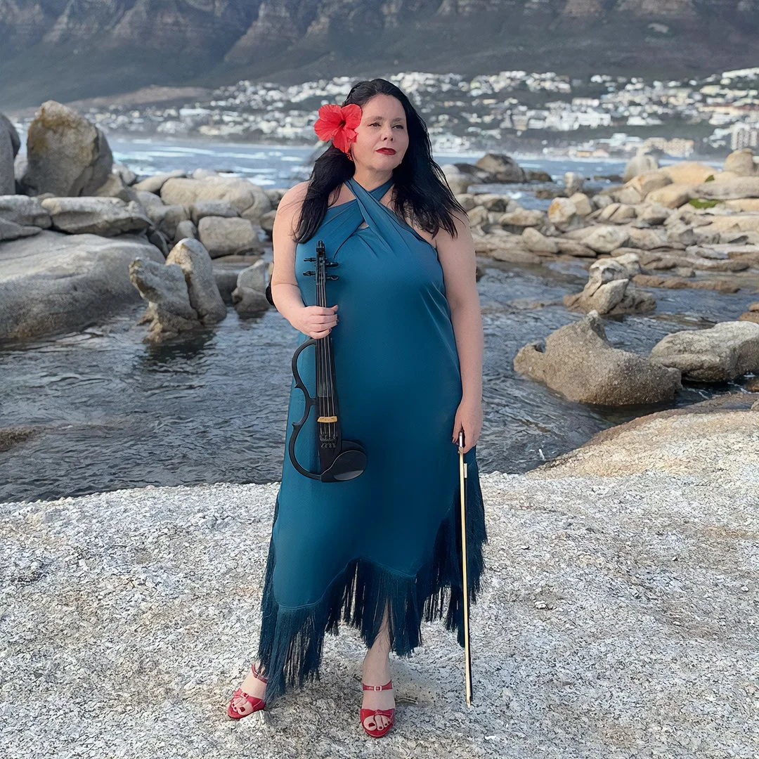 Woman with dark hair, red flower in her hair, wearing a blue dress with fringe, holding a violin and bow, standing on rocky beach with water and mountains in the background.
