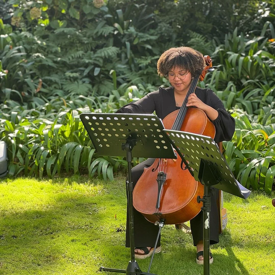Cape Town cello outdoors surrounded by green plants, with music stands in front of her, in bright sunlight.