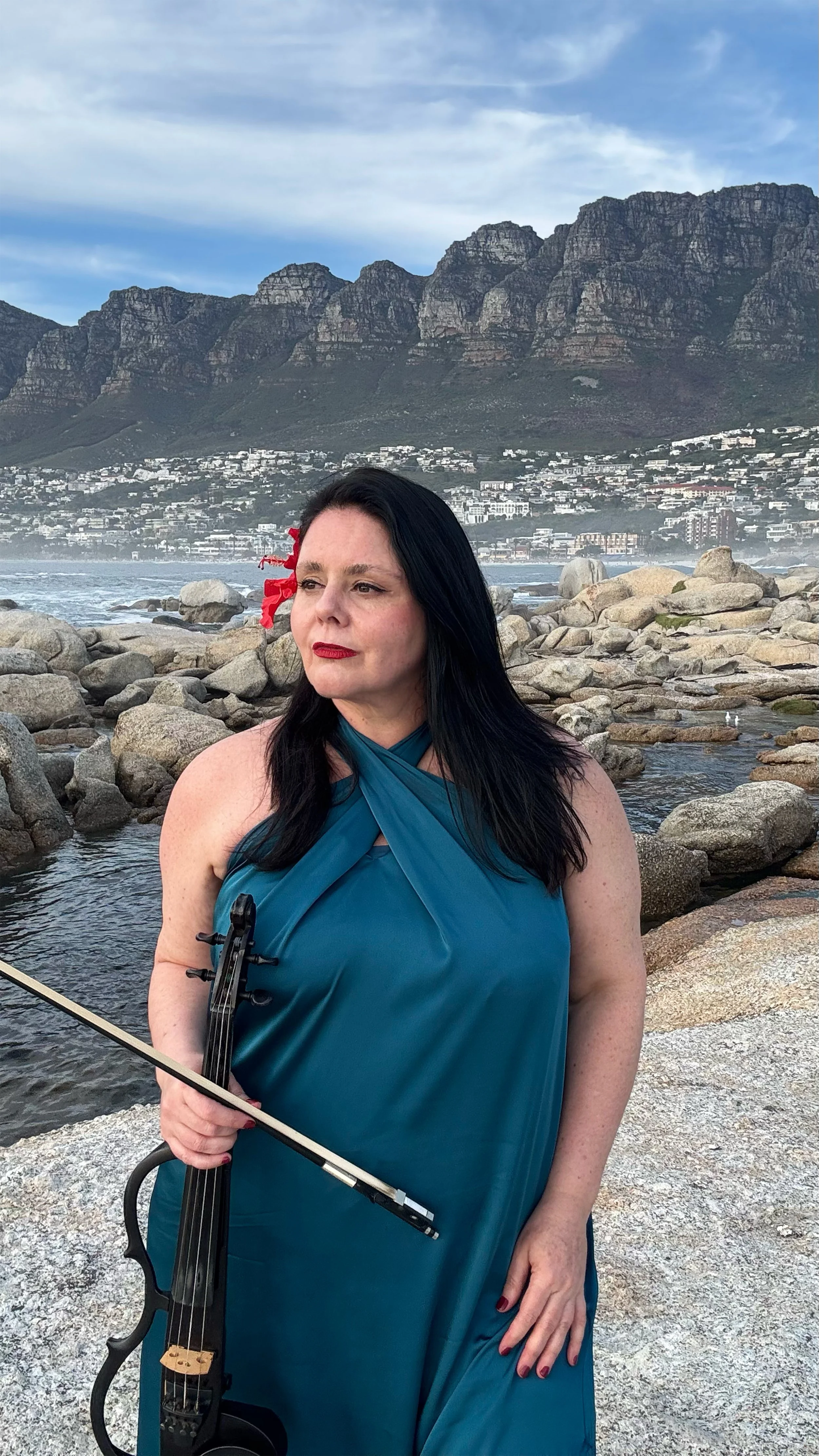 Woman with long dark hair in a teal dress holding a violin on a rocky beach with mountains and a coastal town in the background.