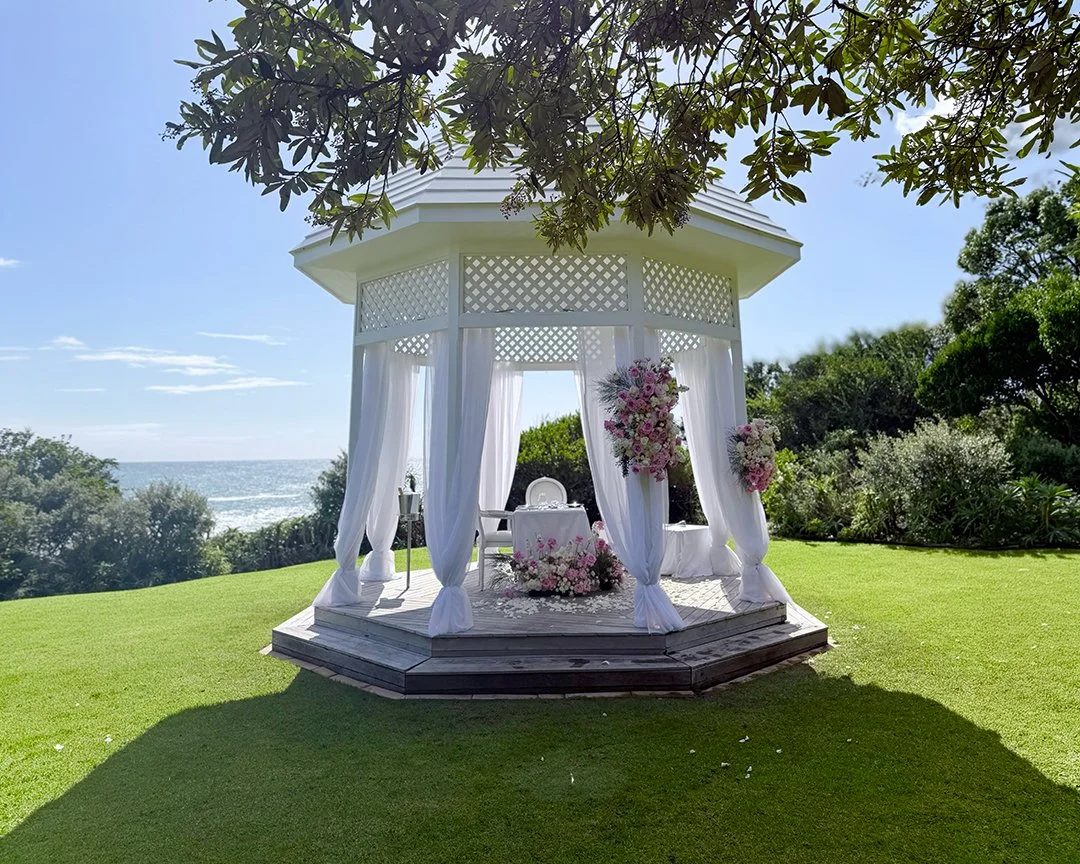 A white wedding gazebo decorated with pink and white flowers, set on a grassy lawn near the ocean at Twelve Apostles Hotel