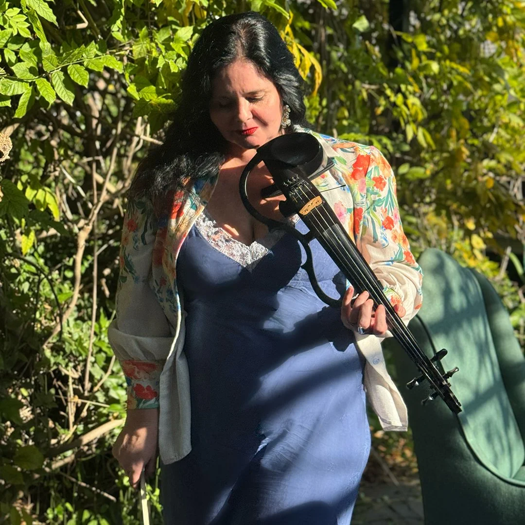 Woman with dark hair playing a violin outdoors surrounded by lush green foliage, wearing a blue dress and a colorful floral jacket.