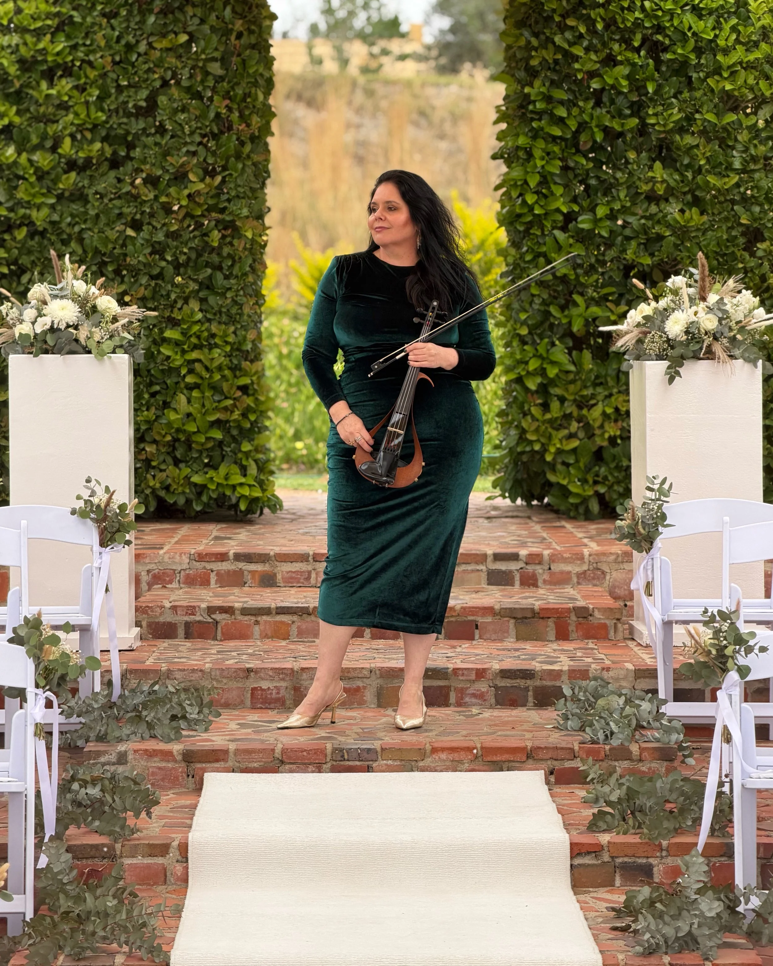 Violinist Larah Eksteen standing in a green velvet dress at an outdoor wedding ceremony venue surrounded by flowers and greenery.
