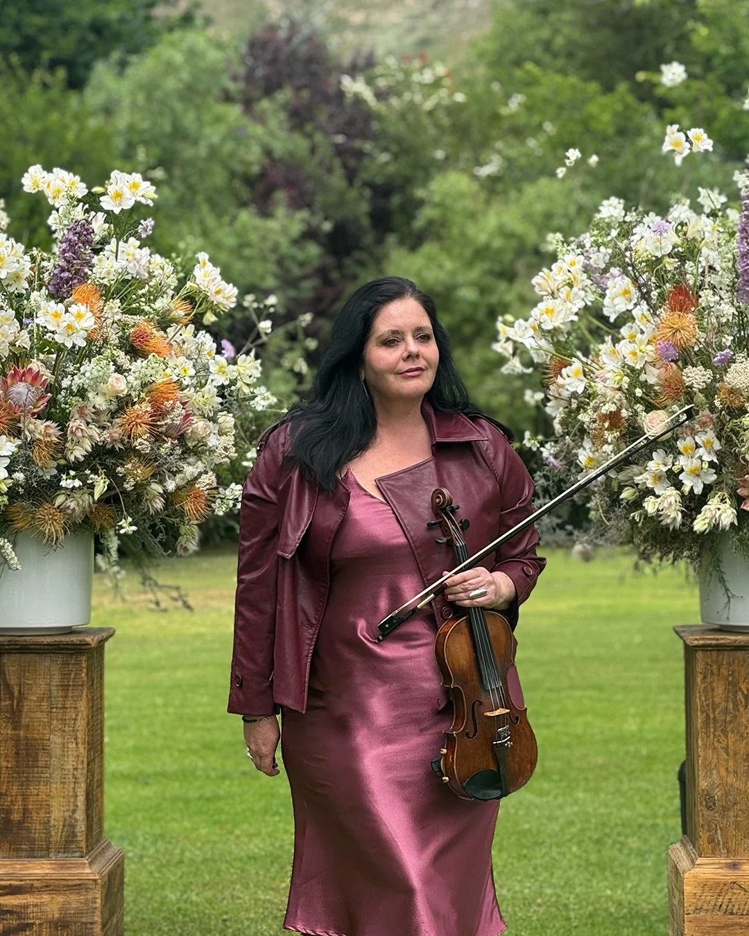 A woman with black hair in a maroon dress and jacket holding a violin and bow, standing outdoors between two large flower arrangements in white pots, with a green background.