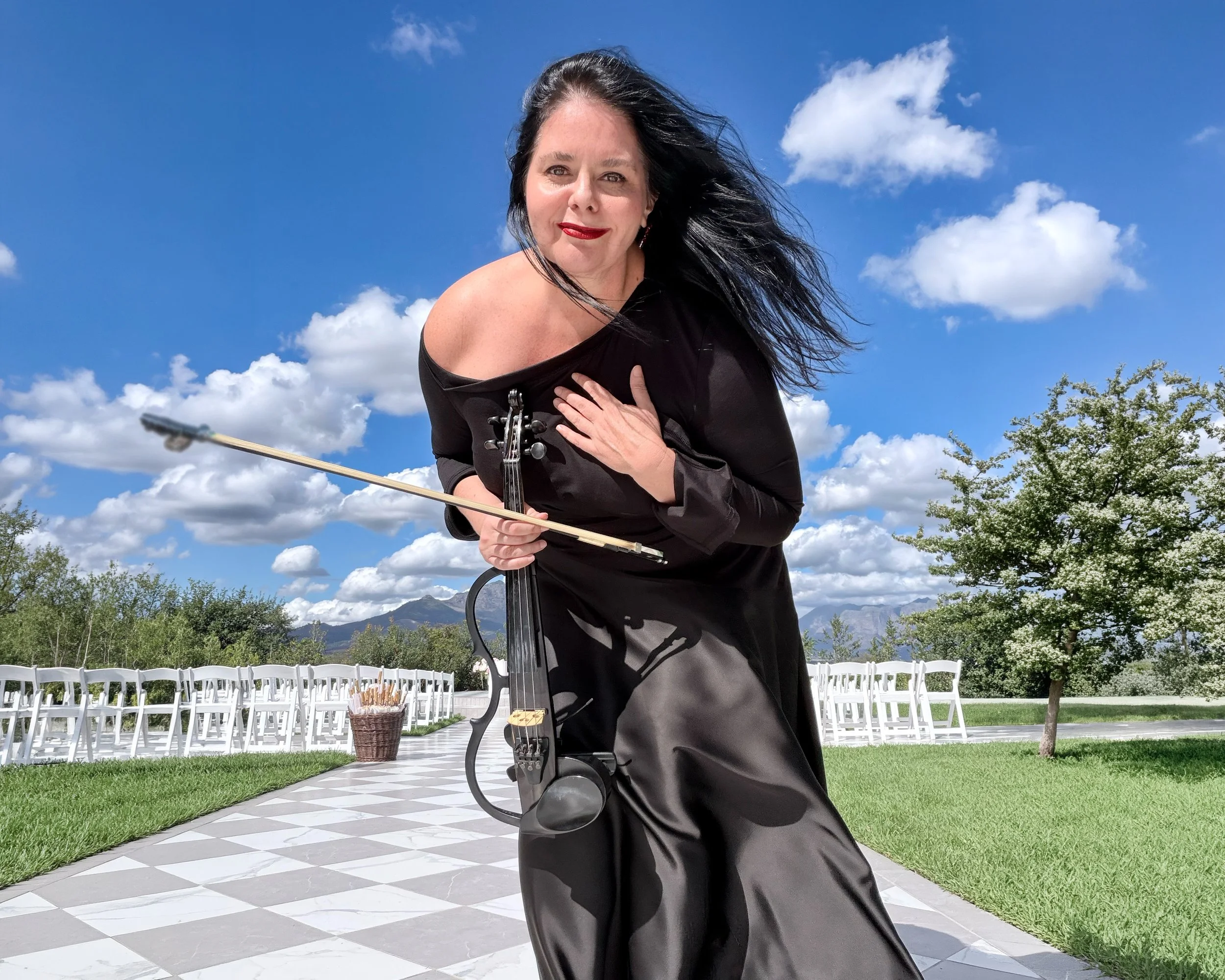 Larah Eksteen at La Paris holding a violin, standing outdoors on a checkered patio with white chairs, a basket of sunflowers, green grass, trees, mountains, and a partly cloudy sky in the background.