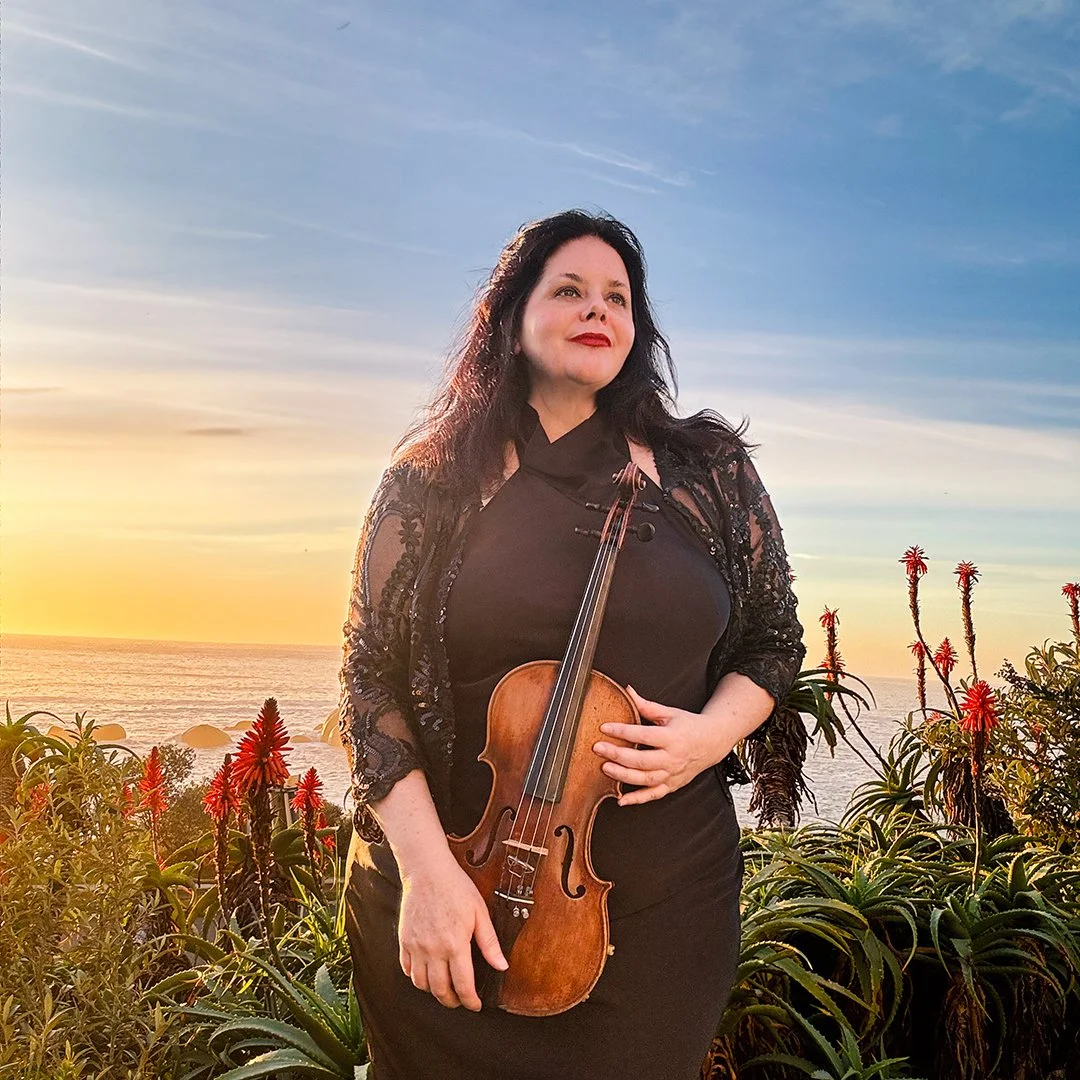 A woman with black hair holding a violin stands outdoors during sunset, with ocean, sky, and plants featuring red flowers in the background.