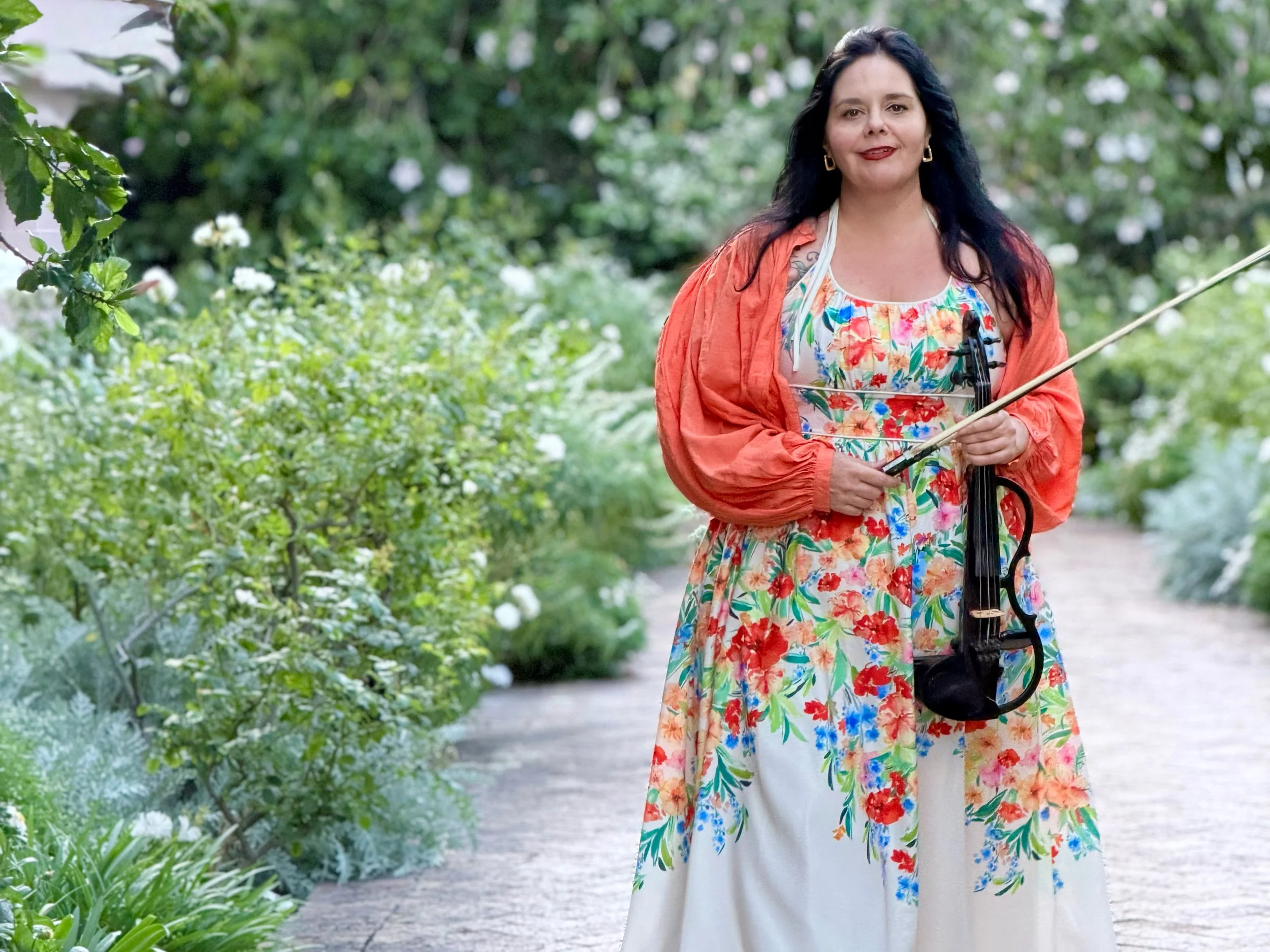 A woman with dark hair, wearing a colorful floral dress and an orange jacket, holding a violin and bow, standing on a garden path surrounded by green foliage and white flowers.