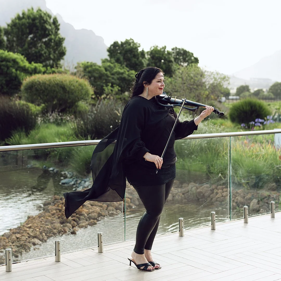 A woman in black clothing playing a violin outdoors on a balcony with a glass railing, overlooking a garden with trees and water.