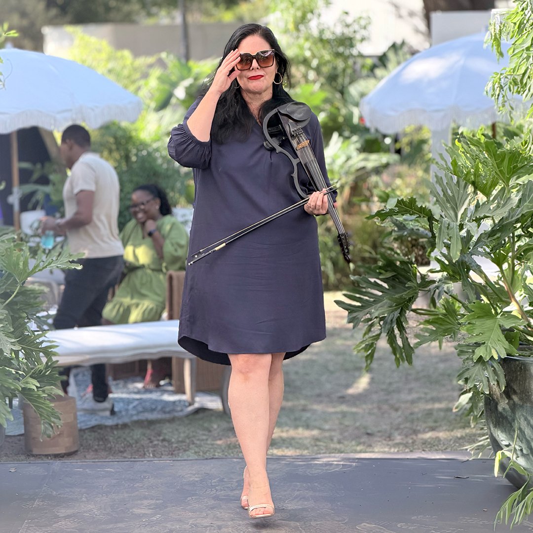 Woman in a black dress and sunglasses holding a violin and bow, standing outdoors at a garden party or event.