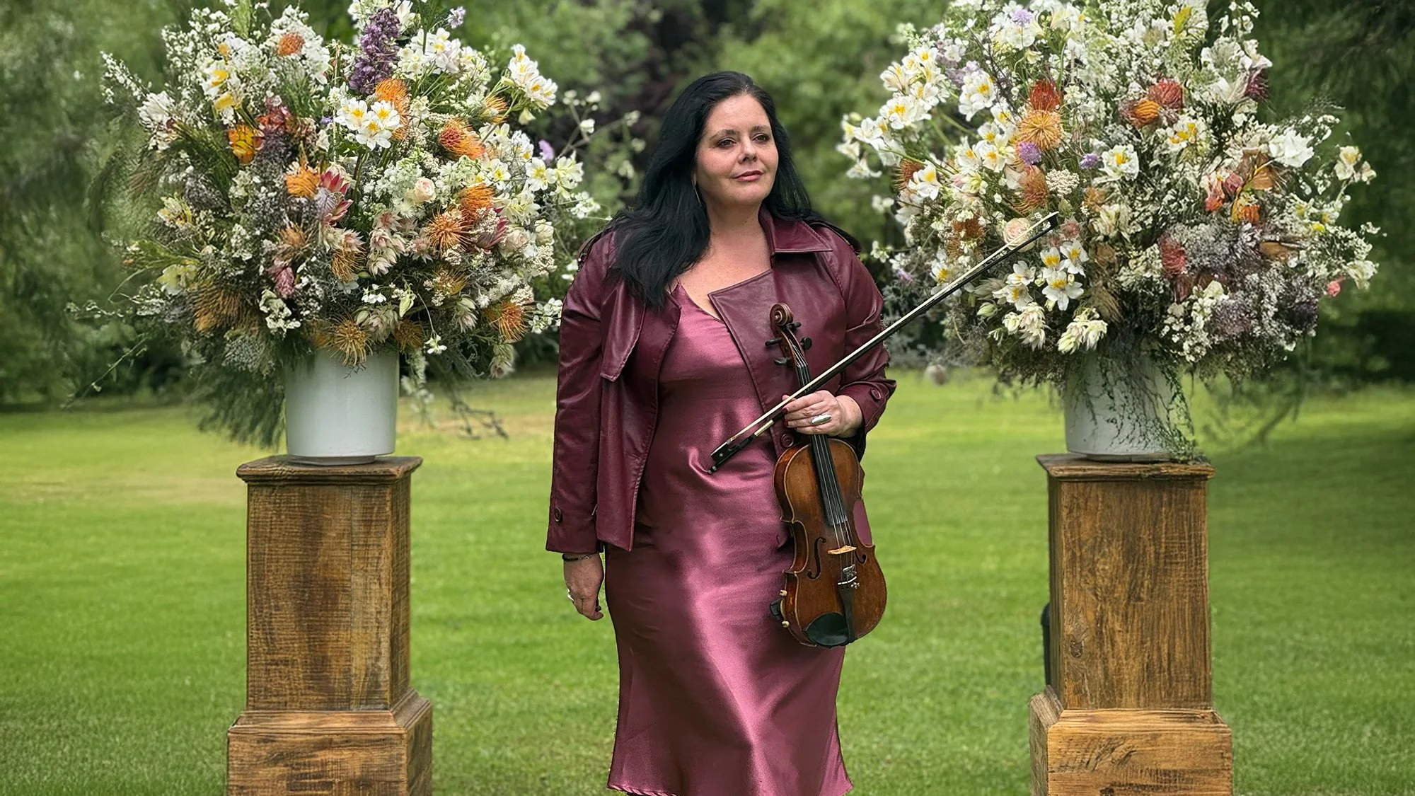 Wedding violinist Larah Eksteen performing at Lanzerac Wine Estate, Stellenbosch — standing between floral ceremony arrangements in a mauve silk dress and burgundy leather jacket, holding her violin