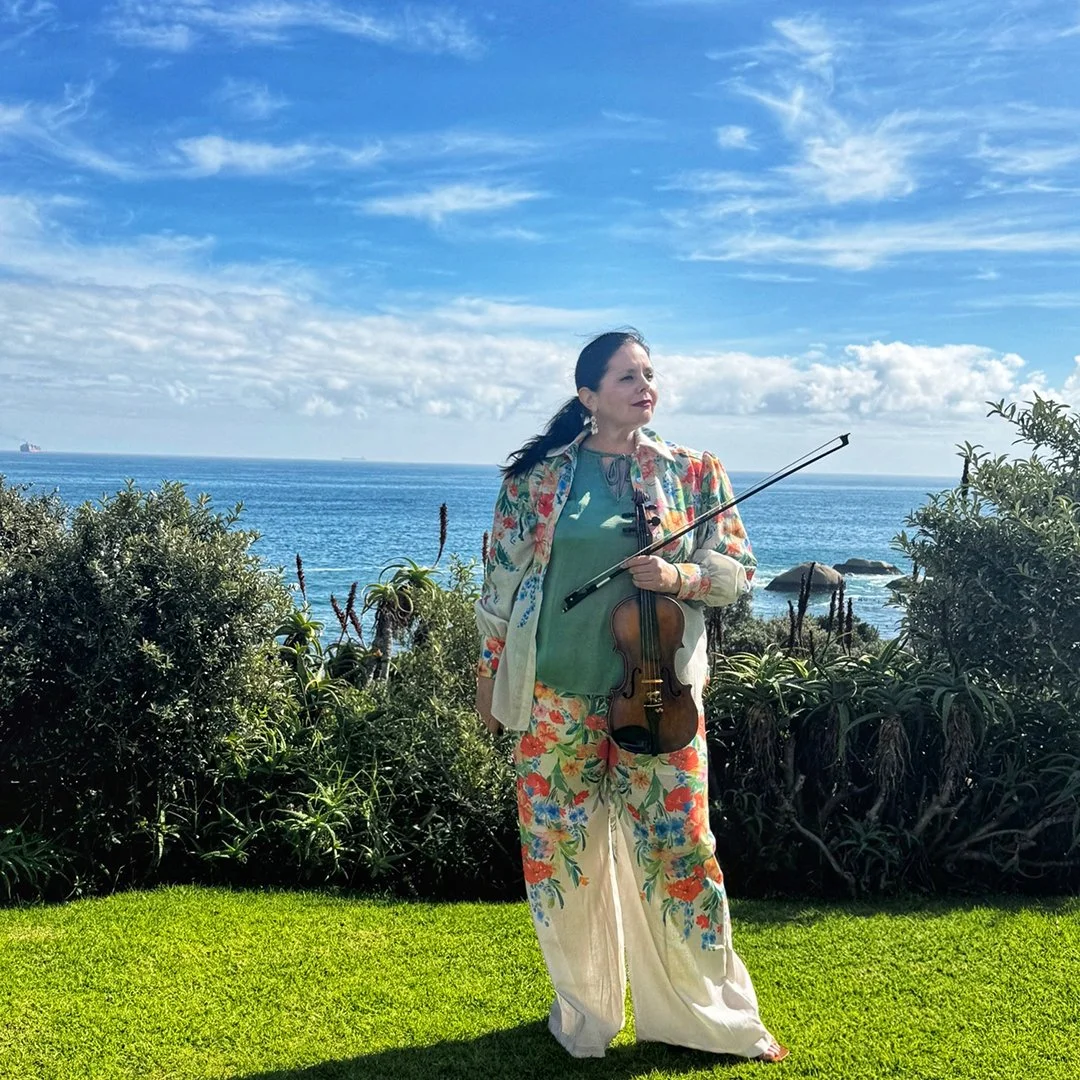 A woman standing outdoors on green grass, holding a violin and a bow, with the ocean and blue sky with clouds in the background.