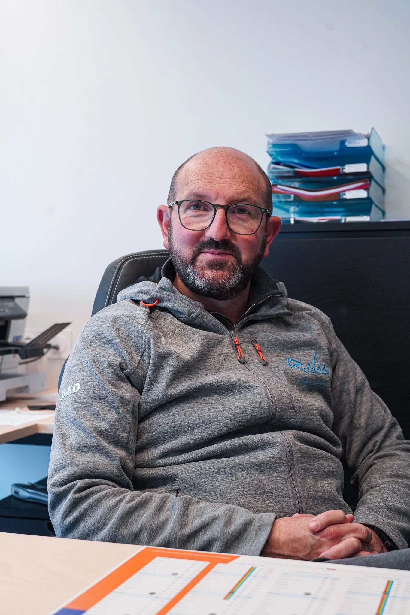 Un homme assis à son bureau, portant un sweat gris, lunettes, dans un bureau avec des piles de dossiers derrière lui.