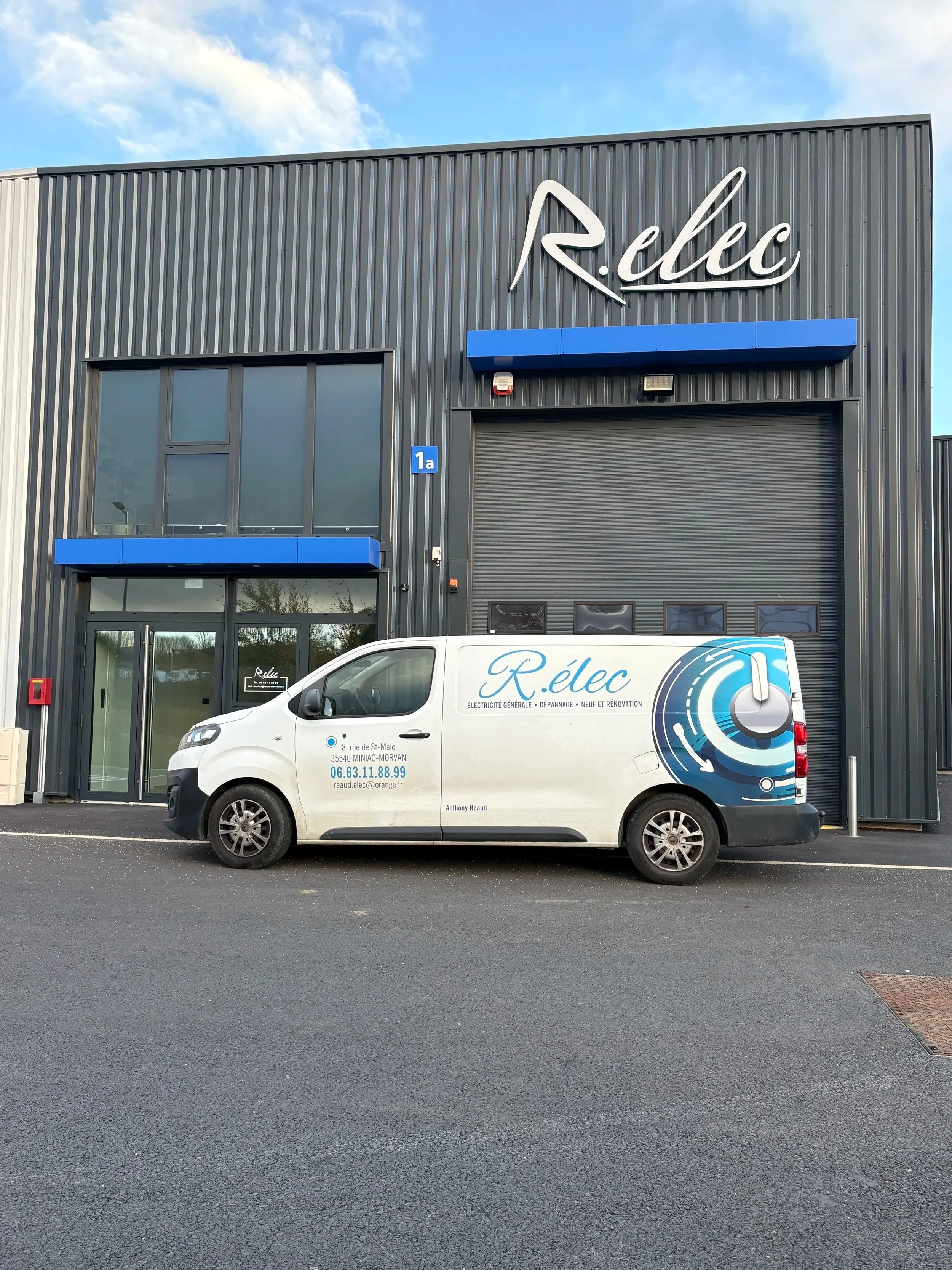 Voiture blanche avec logo et texte 'Rélec, Électricité Générale, Dépannage, Neuf et Rénovation' garée devant un bâtiment industriel gris avec panneaux bleus et un grand logo 'Rélec' sur la façade.