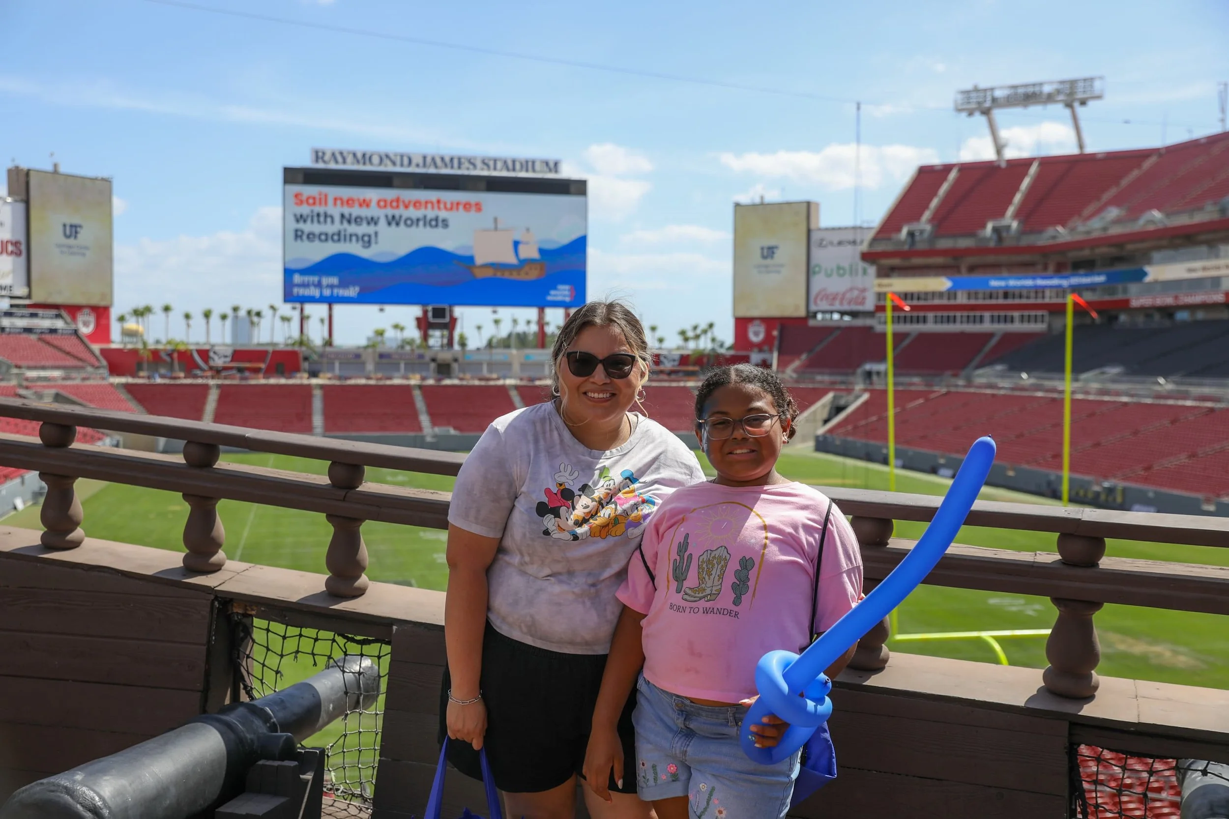 A woman and a girl standing inside Raymond James Stadium with football field in the background. The woman is wearing sunglasses and a Mickey Mouse t-shirt. The girl is wearing glasses and a pink t-shirt, holding a blue balloon twisted into a sword.