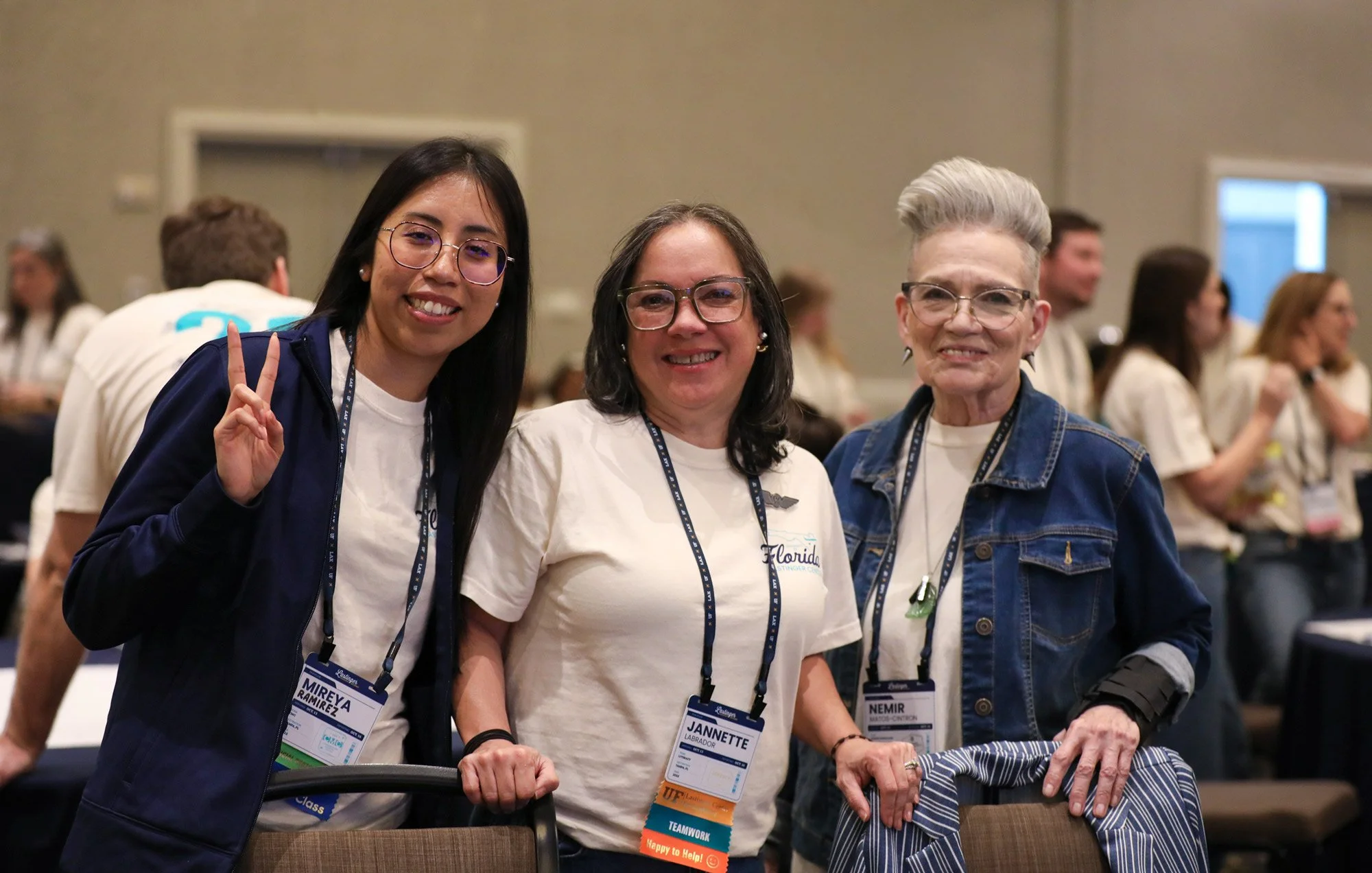 Three women standing together at a conference, smiling at the camera. The woman on the left is making a peace sign. All are wearing conference badges around their necks.