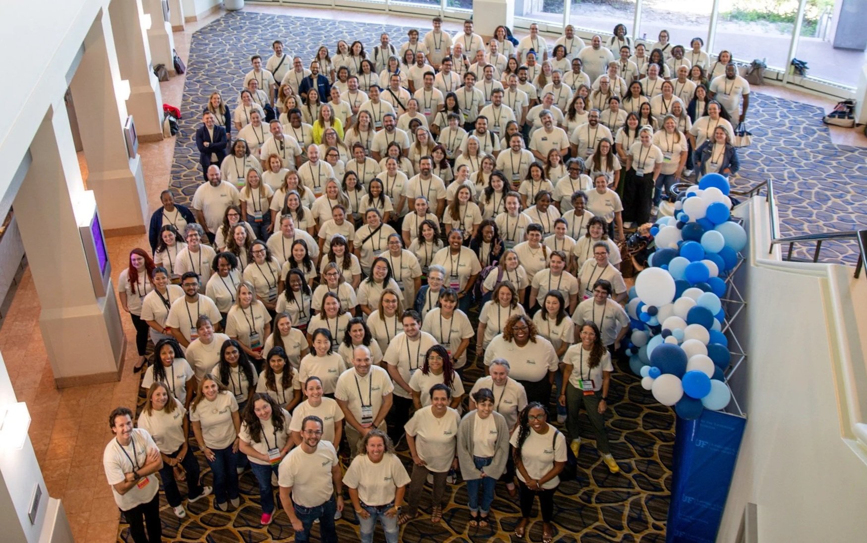A large group of people gathered in a conference hall for a group photo. They are wearing matching white shirts and standing on patterned carpet near a display of blue and white balloons.