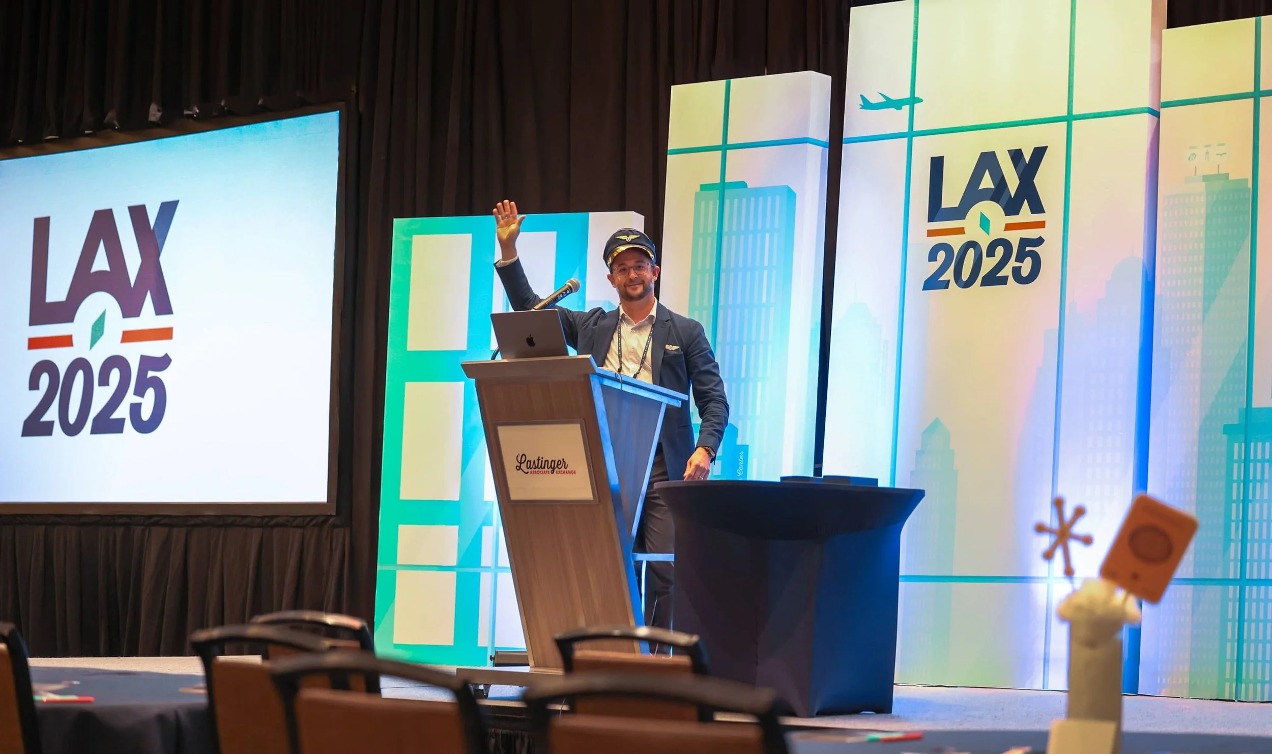 A man in a pilot costume stands at a podium on a stage, waving and smiling. The stage has a backdrop displaying the Arrivals Lounge Exchange LAX 2025 logo with cityscape graphics, and a large screen showing the same logo. There are tables and chairs in the foreground, and a small table with a decorative item on it to the right.