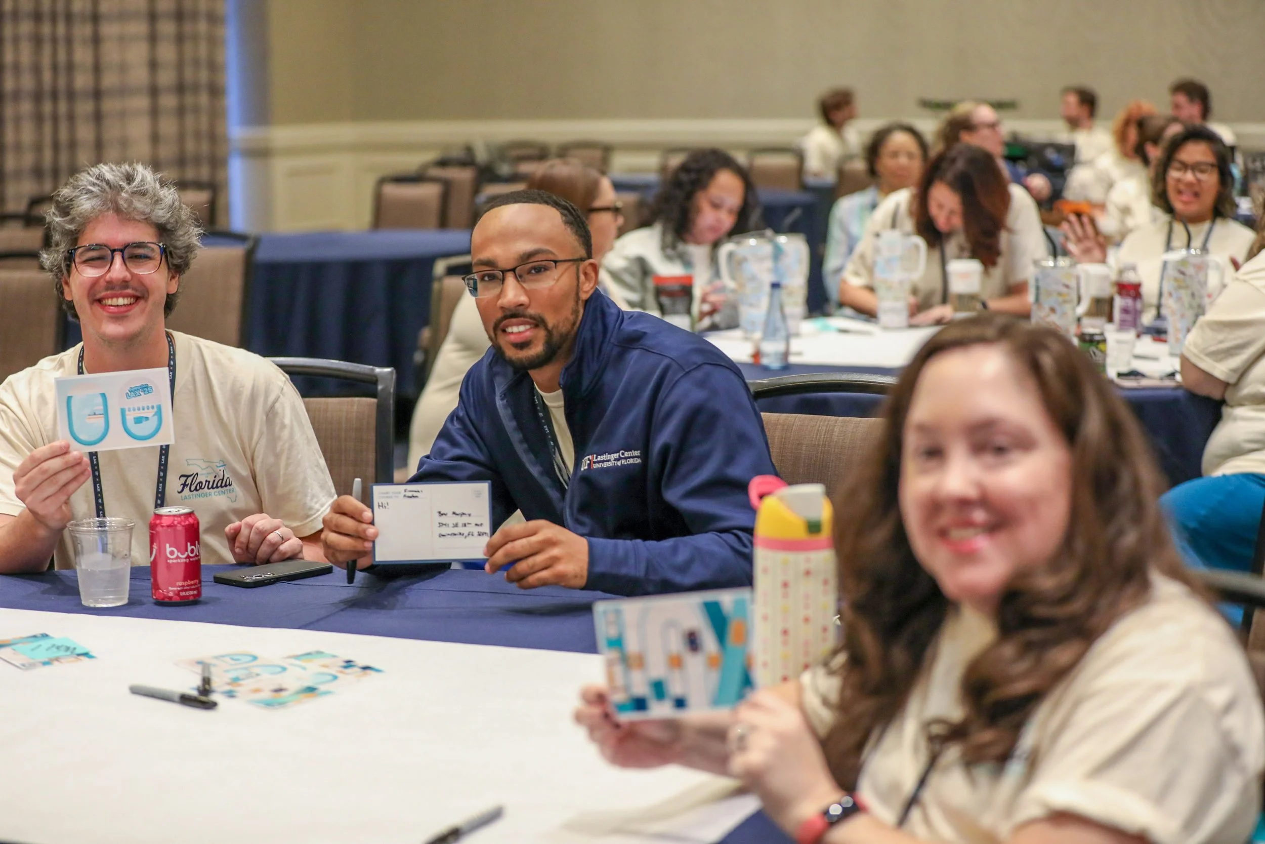 Group of people at a conference or workshop, sitting at tables with notebooks, drinks, and name tags, engaging in activities and smiling