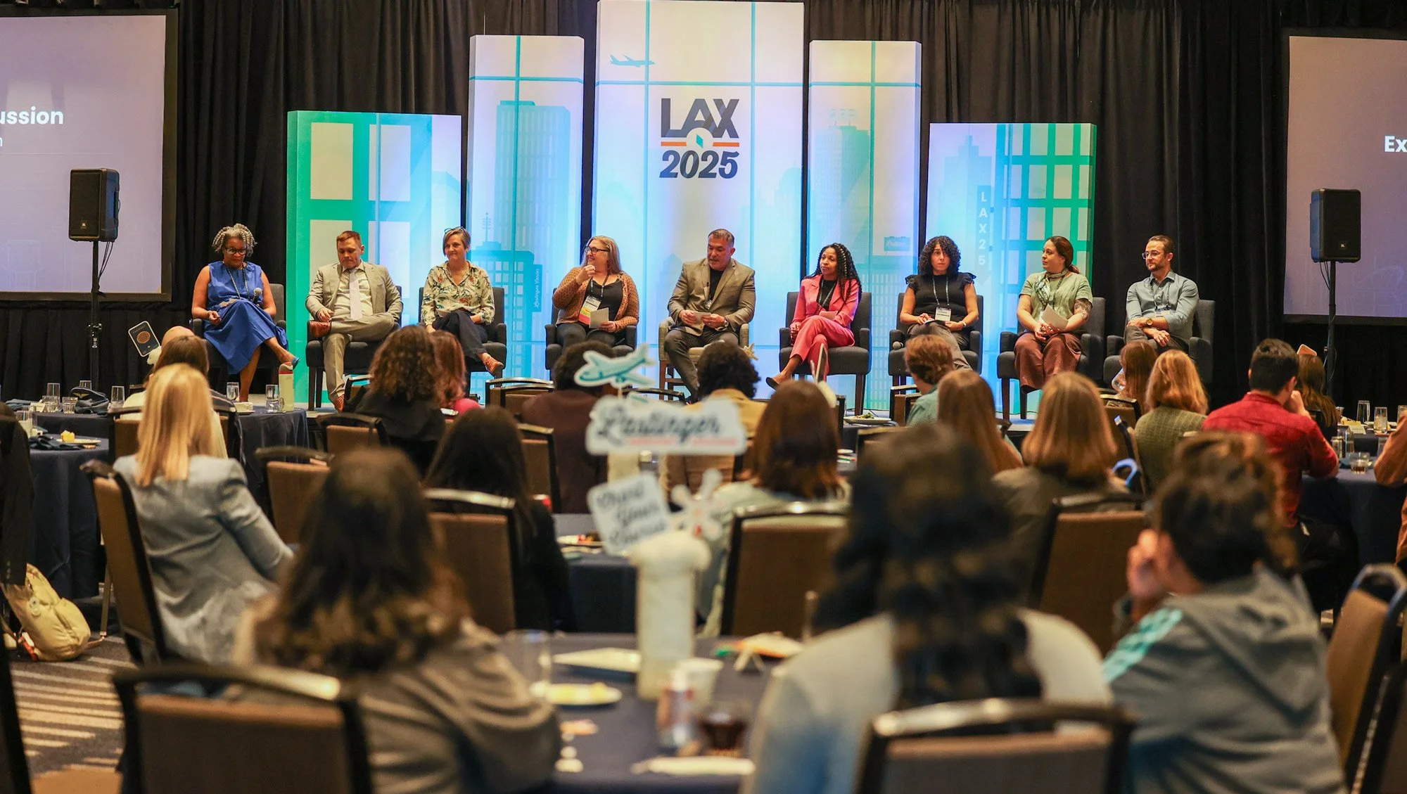 A panel of nine people sitting on chairs on stage during a conference, with an audience seated at round tables in front. The stage backdrop displays the LAX 2025 logo and cityscape graphics, and there's a large screen on each side of the stage.