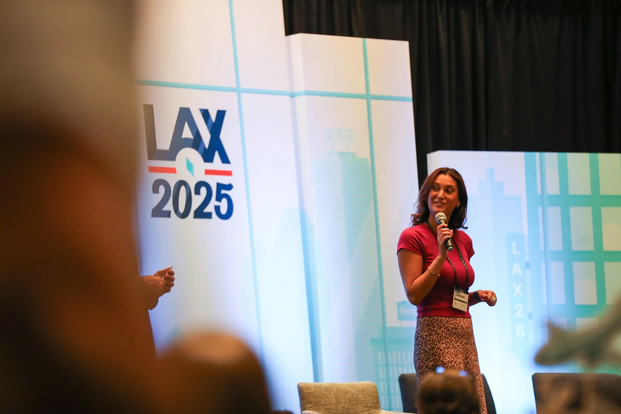 A woman in a red top and patterned skirt holding a microphone speaking at a conference or event, with a large presentation screen behind her displaying 'LAX 2025' and a cityscape graphic.