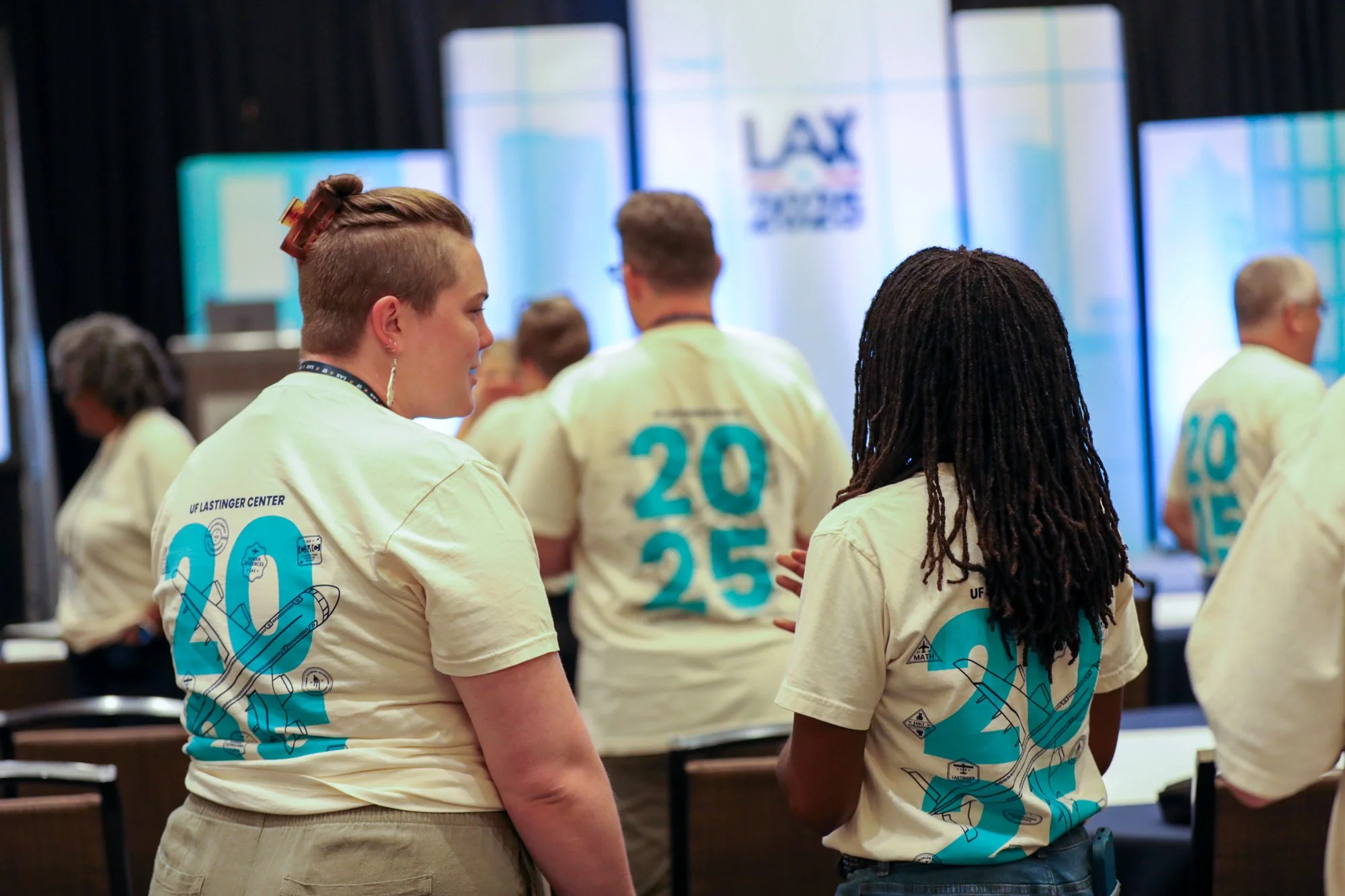 People wearing cream-colored T-shirts with blue graphics and numbers, standing in a conference or event room with screens displaying 'LAX' and '2025' in the background.