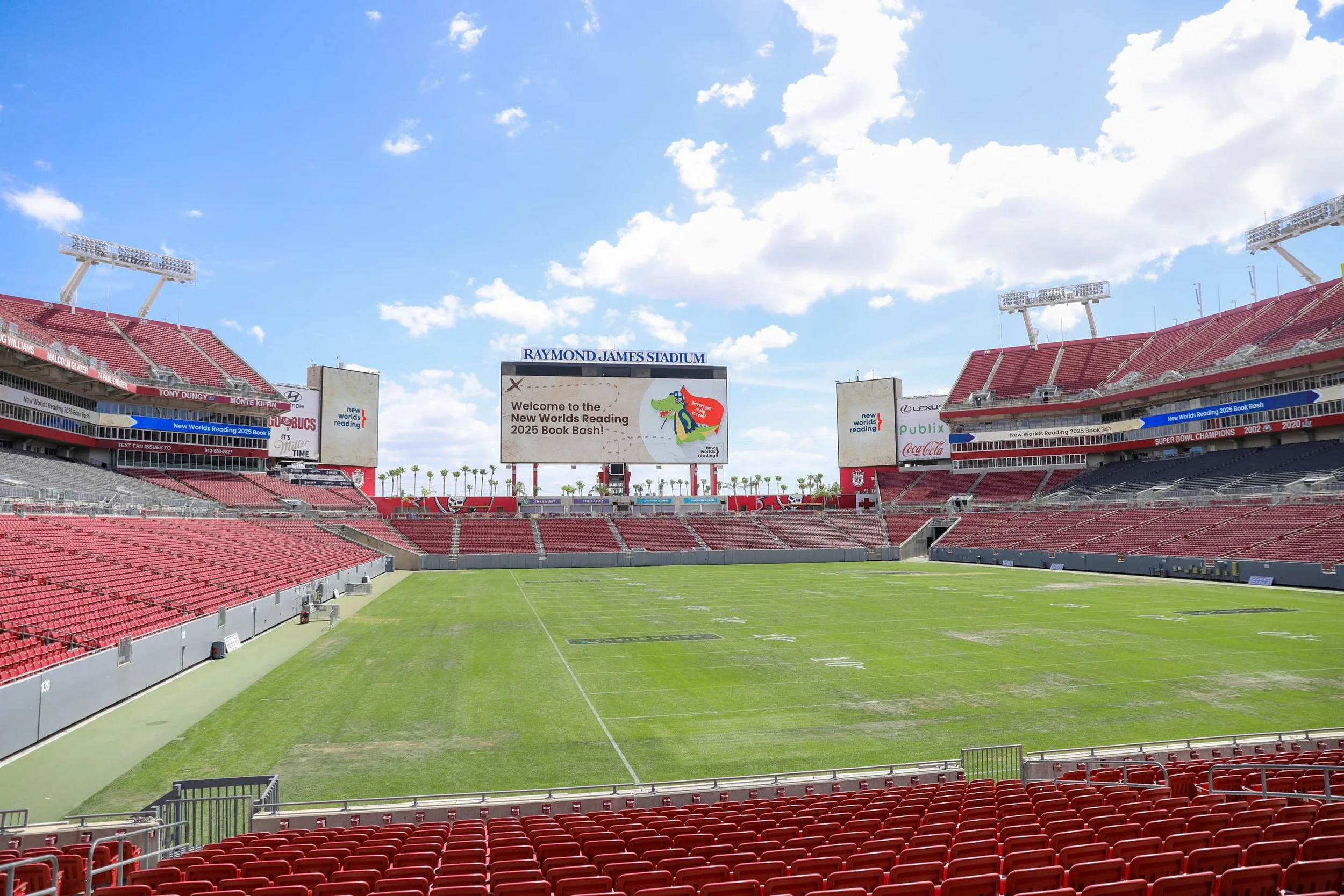 Empty football stadium with red seats, a green field, and a large scoreboard displaying a message about a book event, under a partly cloudy sky.