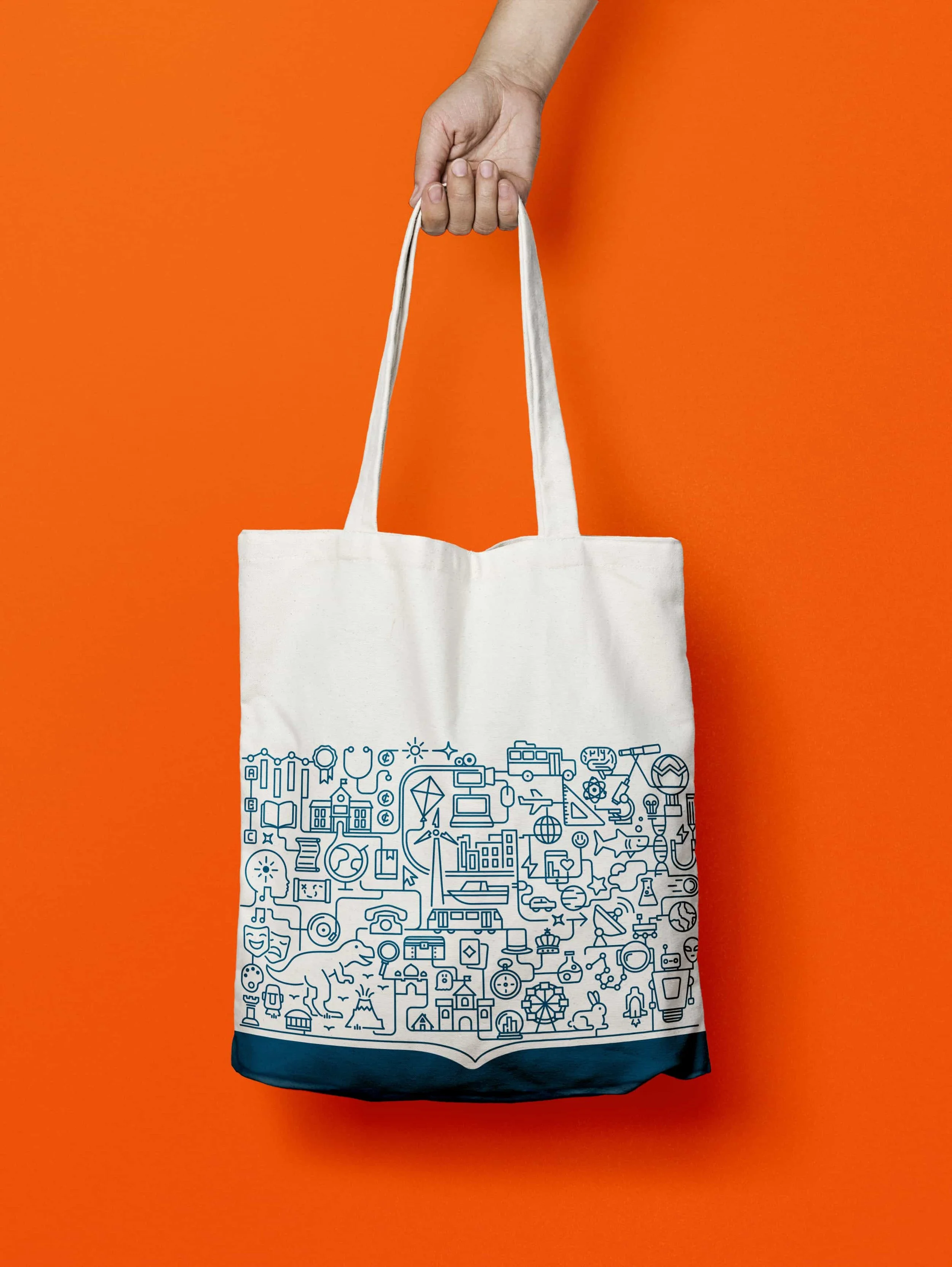 A white tote bag with a blue bottom, held by a hand against an orange background. The bag features a blue line drawing of various icons, including a dinosaur, books, a telescope, and other science and technology-related images.