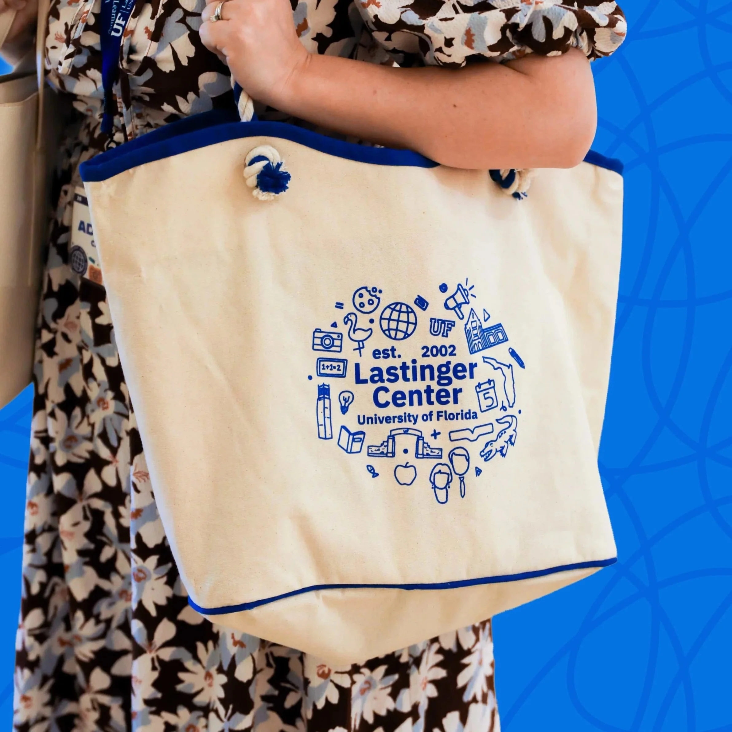 Person holding a cream-colored tote bag with blue trim and print from Lastinger Center at the University of Florida, featuring various educational and university-related icons.