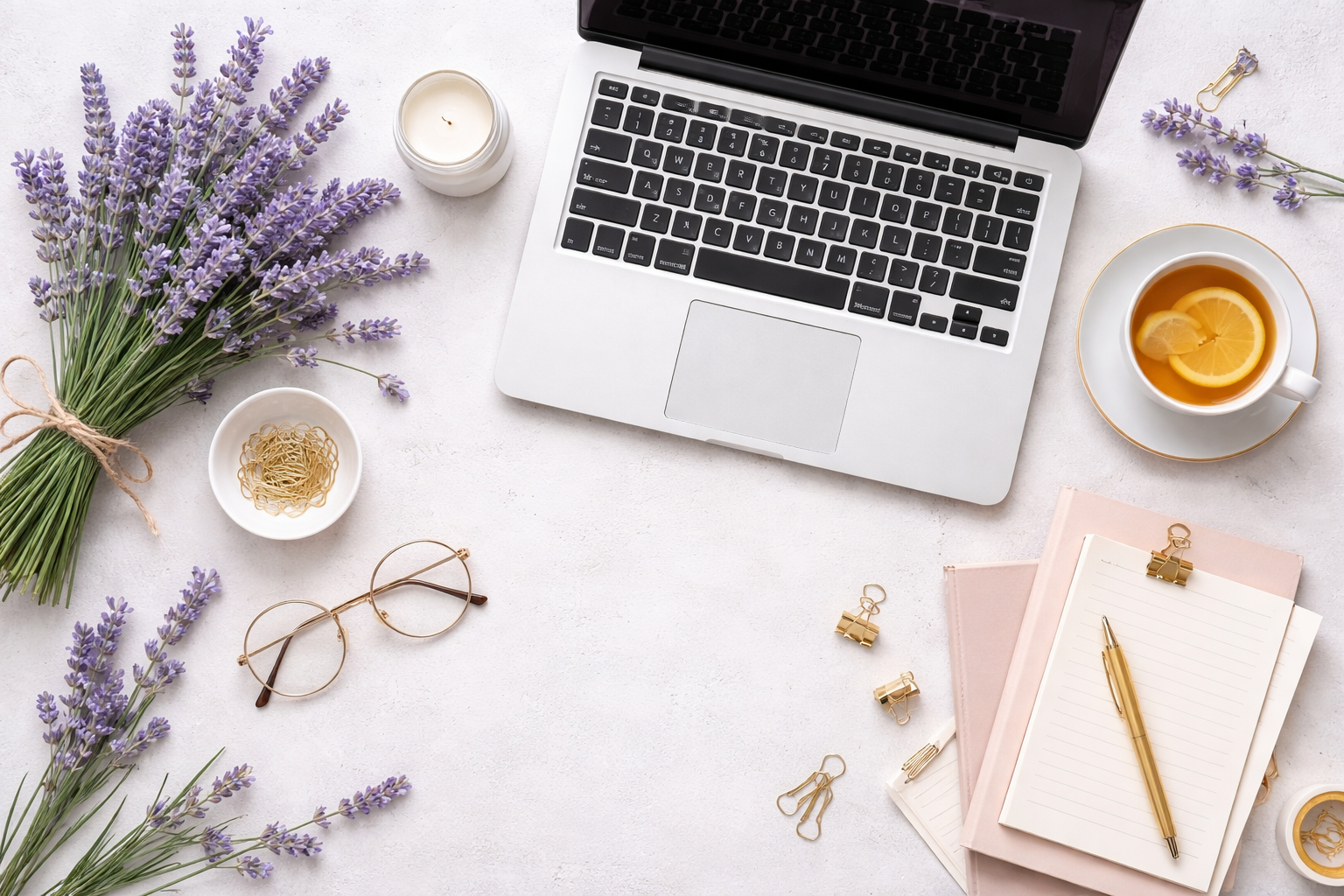A workspace with a laptop, a cup of tea with lemon, a bunch of lavender, glasses, notebooks, pens, and decorative clips on a white surface.