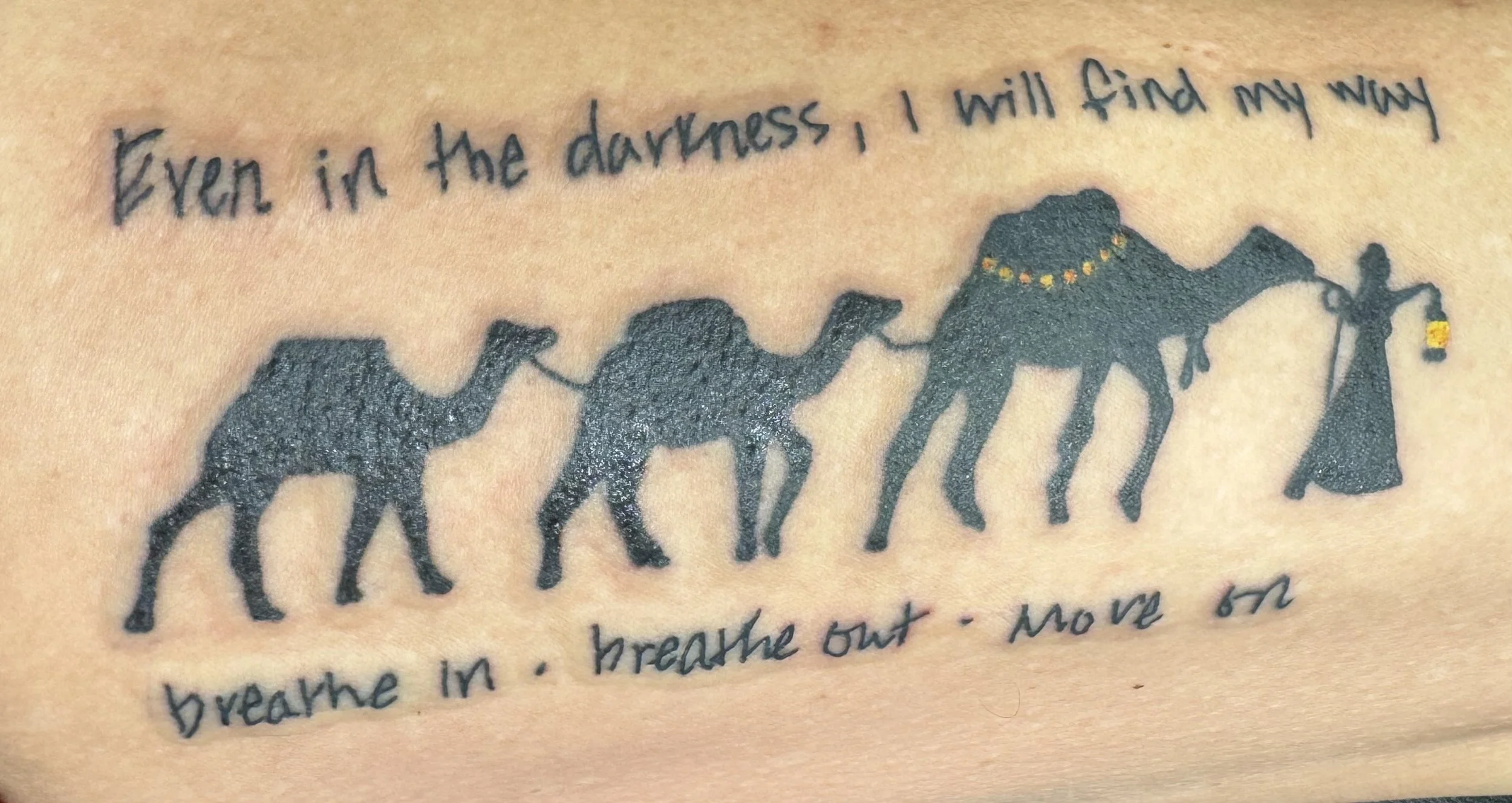 Tattoo of a line of four black camels, each connected by a line, with a handwritten quote above and below. The quote reads: 'Even in the darkness, I will find my way' above, and 'breathe in. breathe out. move on' below.