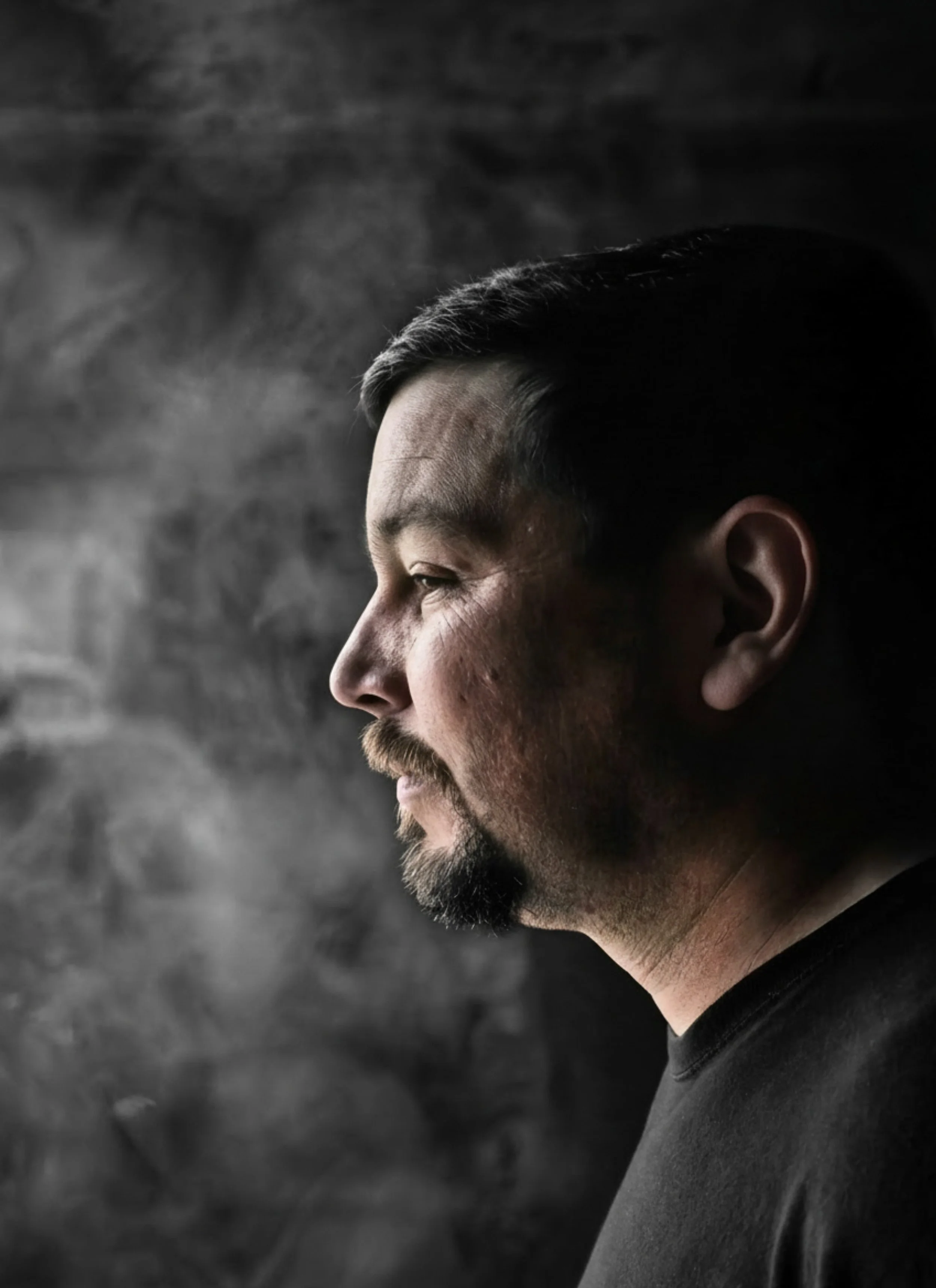 Side profile of a man with short dark hair, beard, and mustache against a dark, textured background.