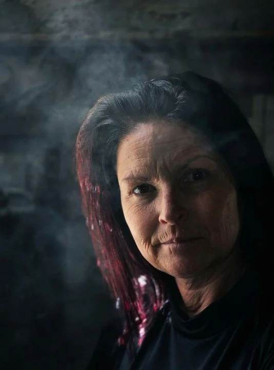 Close-up of a woman with dark hair streaked with red, looking directly at the camera with a serious expression in a smoky, dark background.