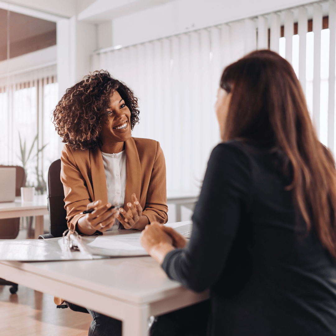 Two women having a conversation in a professional office setting, with one woman smiling and gesturing while the other listens.