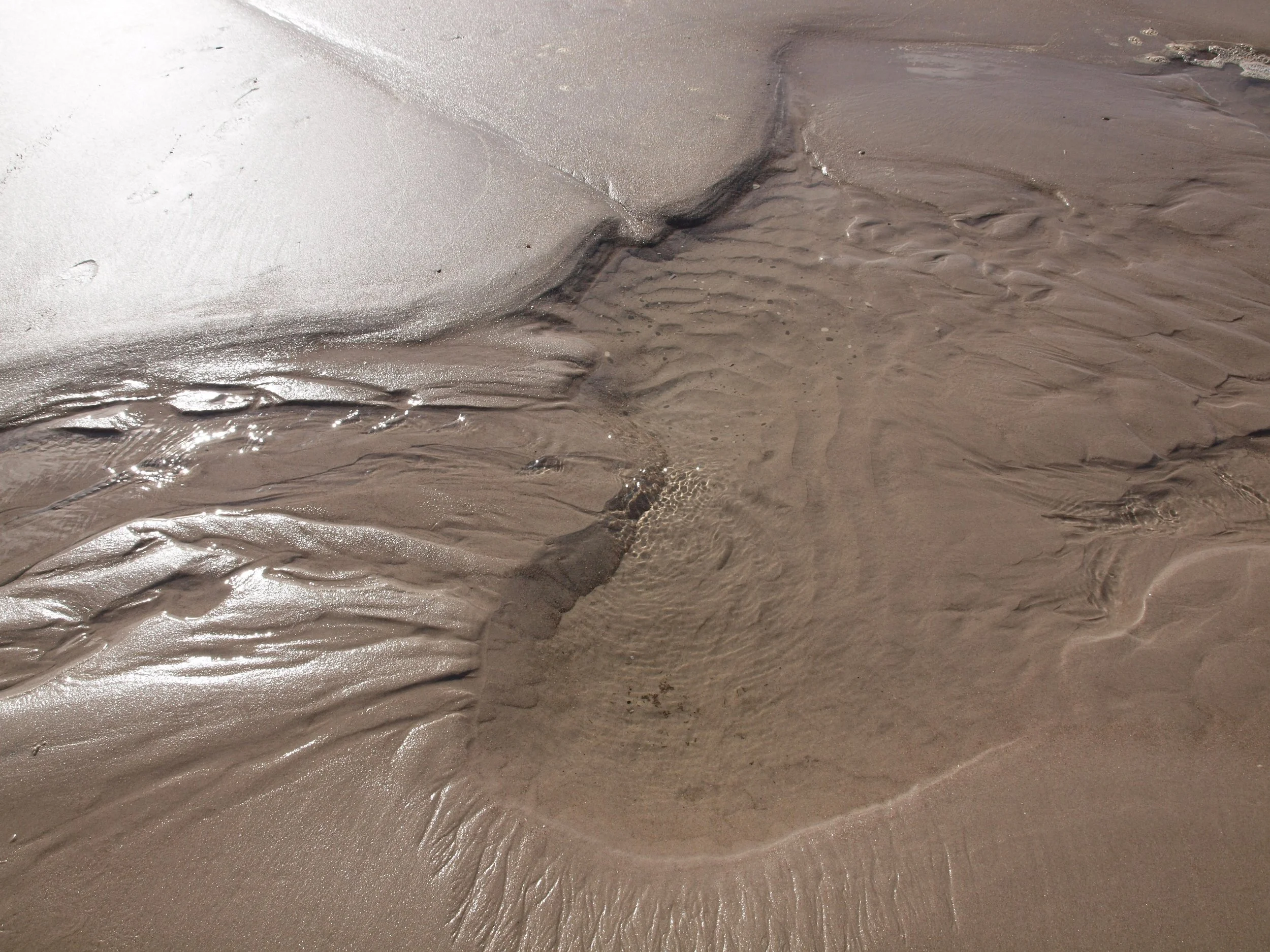 Feuchter Sand der stellenweise mit Wasser durchzogen ist, in dem sich die Sonne spiegelt.