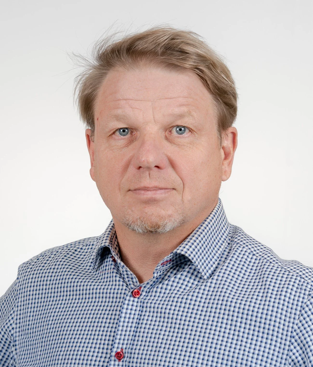 Kuva keskellä ikäisestä miehestä, jolla on vaaleat hiukset ja siniset silmät, pukeutuneena sinisiin ruutupaitaan, mustavalkoisella taustalla.