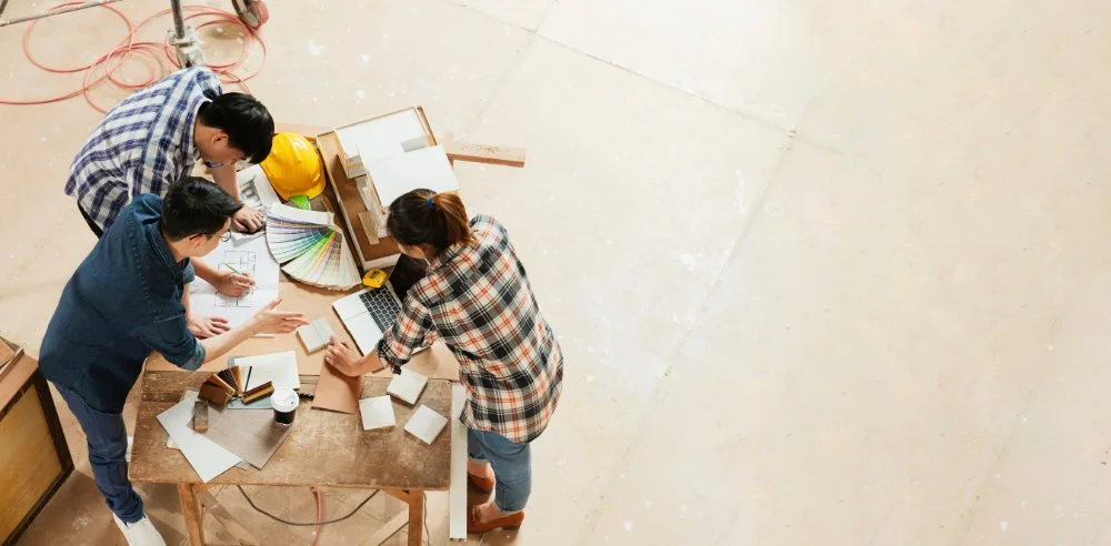 Three people at a worktable review color swatches, papers, and tiles, suggesting a design or construction planning meeting.