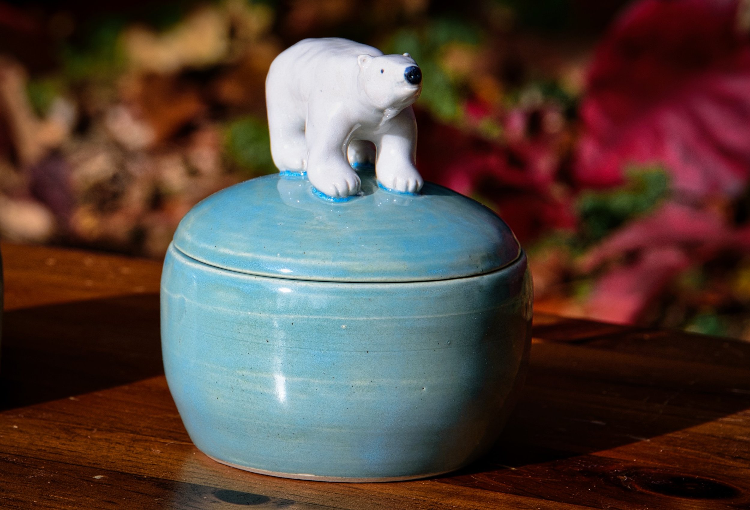 A ceramic jar with a light blue glaze and a bear figurine on top, placed on a wooden surface outdoors with autumn foliage in the background.