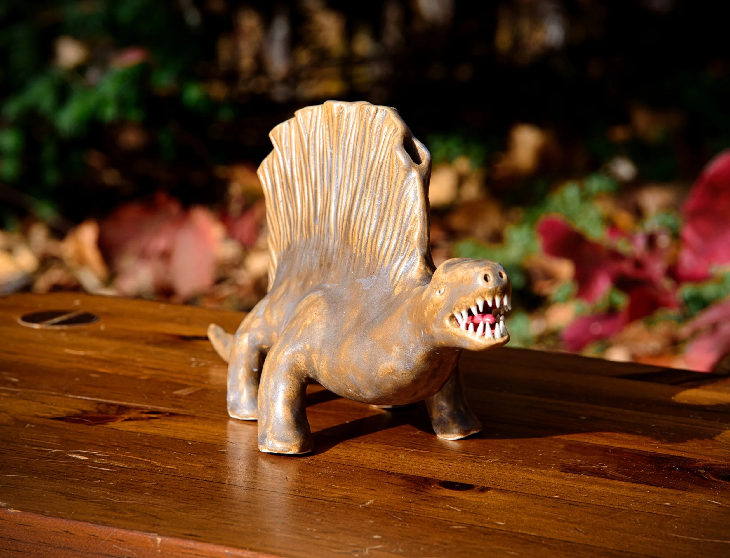 A small figurine of a creature with the body of a horse and the head of a dinosaur, resembling a mythical or fictional animal, is placed on a wooden surface outdoors with blurred autumn leaves in the background.